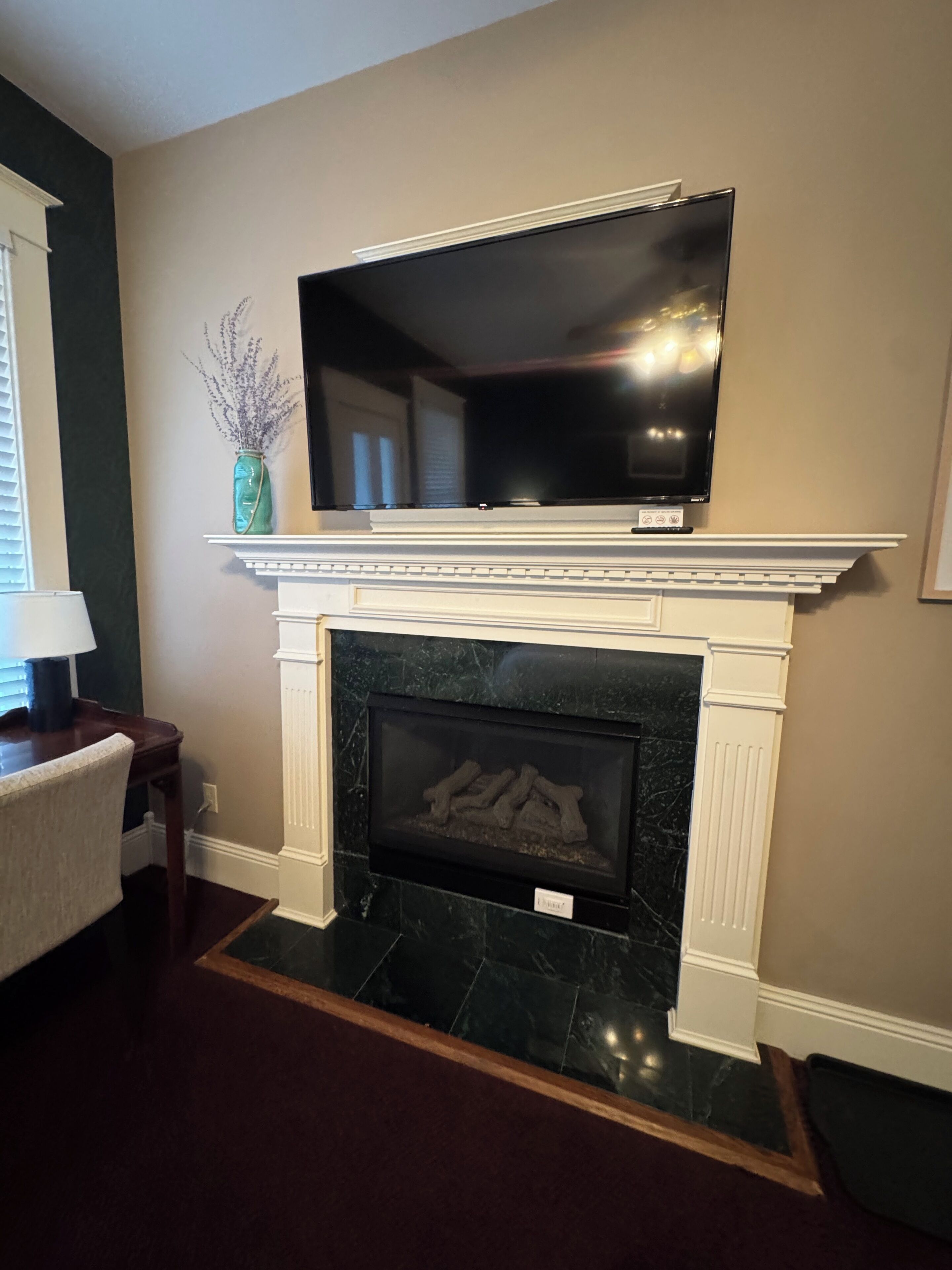 Una sala de estar con chimenea, un televisor montado encima, un jarrón con flores secas y una lámpara en una mesita auxiliar.