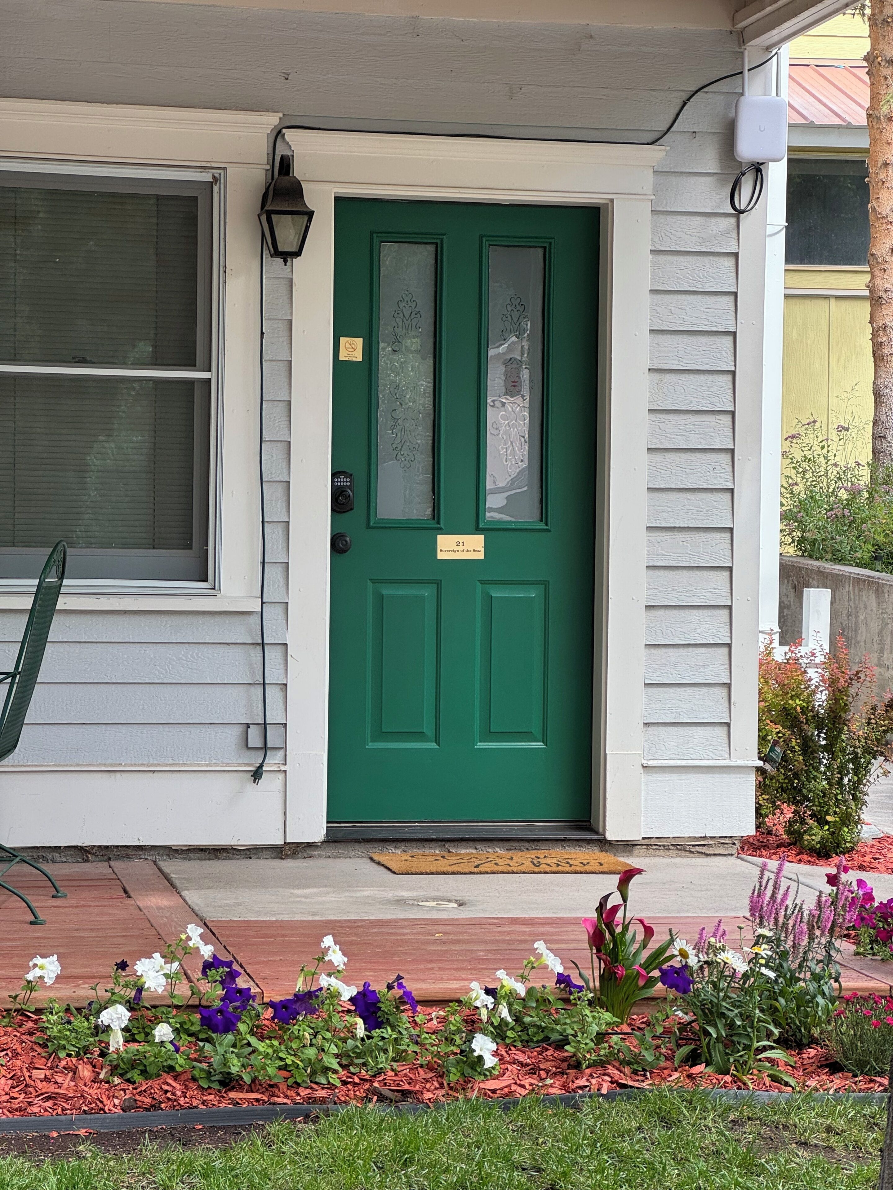 A green door with a number plate, a window with blinds, and a small porch area with a chair and flower beds.