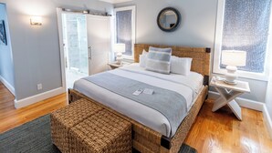 A modern bedroom with a large bed, a wicker headboard, a bedside table with a lamp, and a view of the outside through a window. - Town & Tide Inn (Newport)