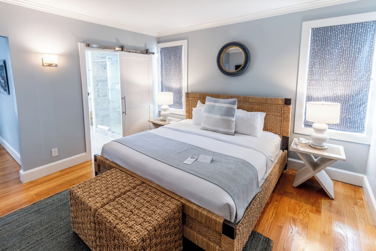 A modern bedroom with a large bed, a wicker headboard, a bedside table with a lamp, and a view of the outside through a window.