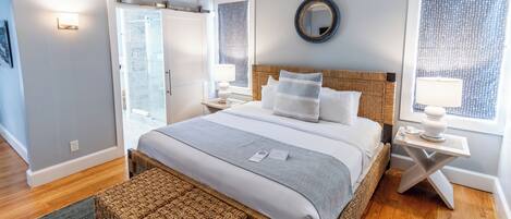 A modern bedroom with a large bed, a wicker headboard, a bedside table with a lamp, and a view of the outside through a window.