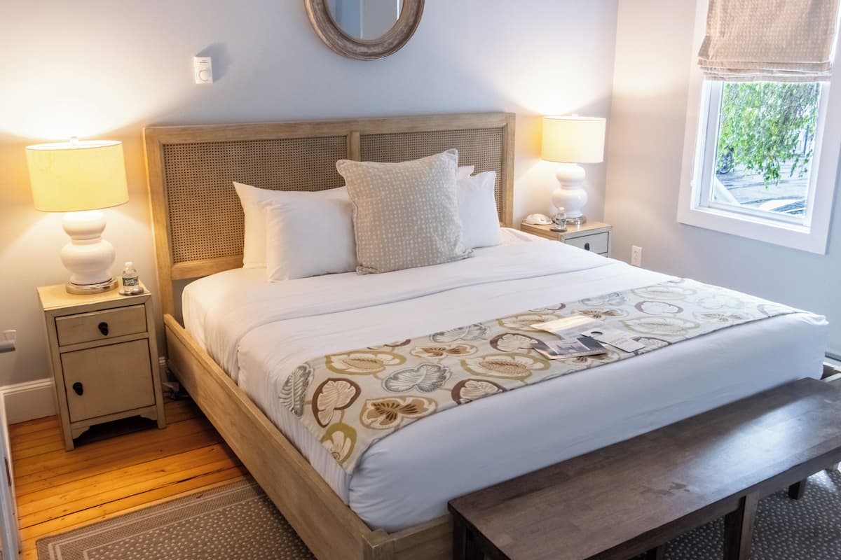 A neatly made bed with a floral-patterned blanket, two bedside lamps, a wooden headboard, and a window with a view of greenery.