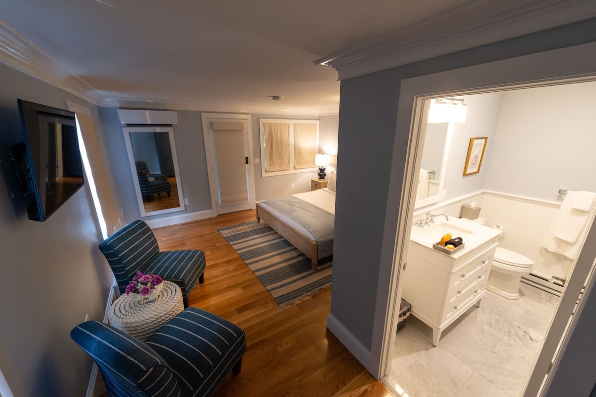A modern bedroom with a bed, a sitting area with a chair and ottoman, a bathroom with a sink and toilet, and a hallway leading to another room.