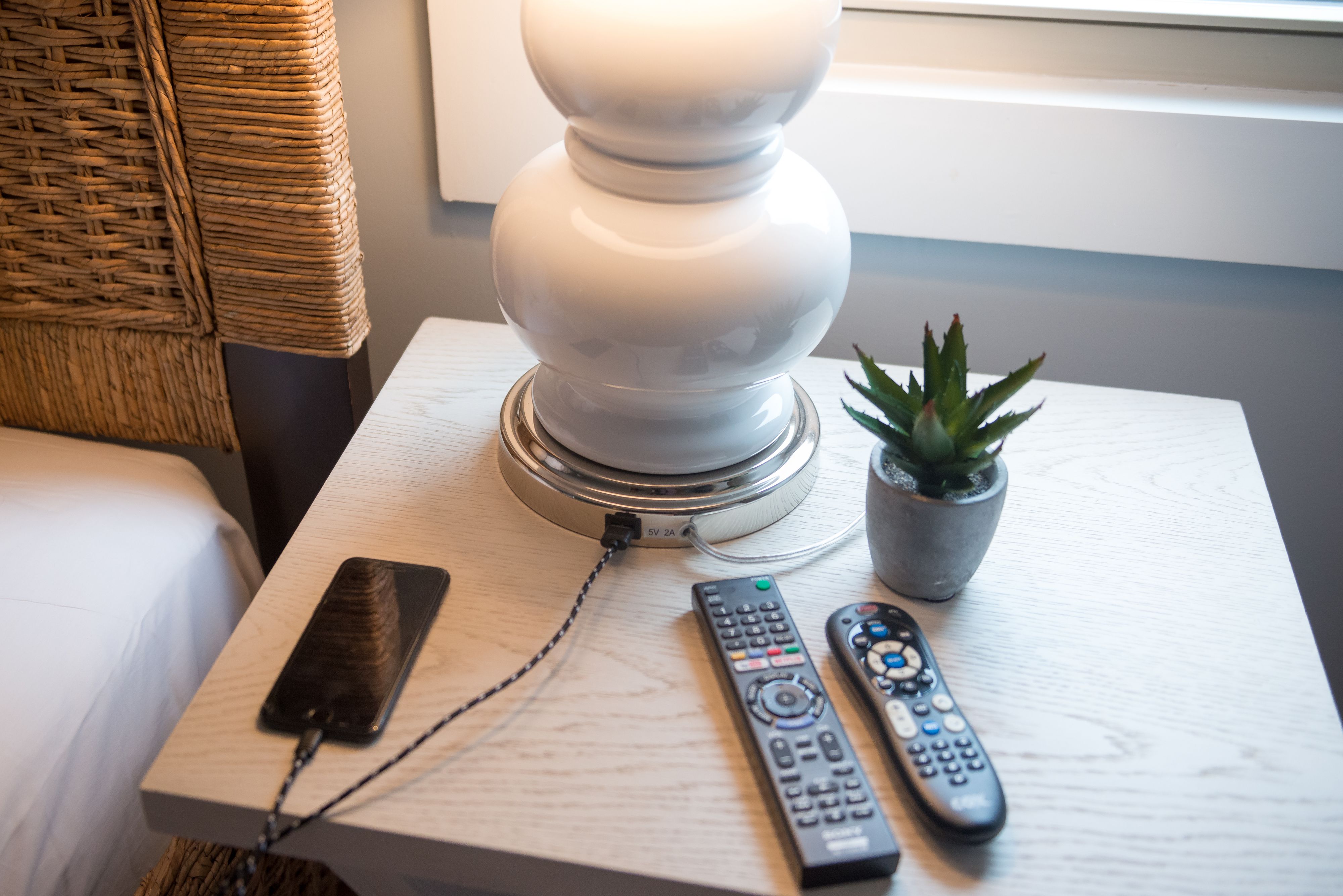 Uma mesa de cabeceira com uma luminária, um smartphone e dois controles remotos.