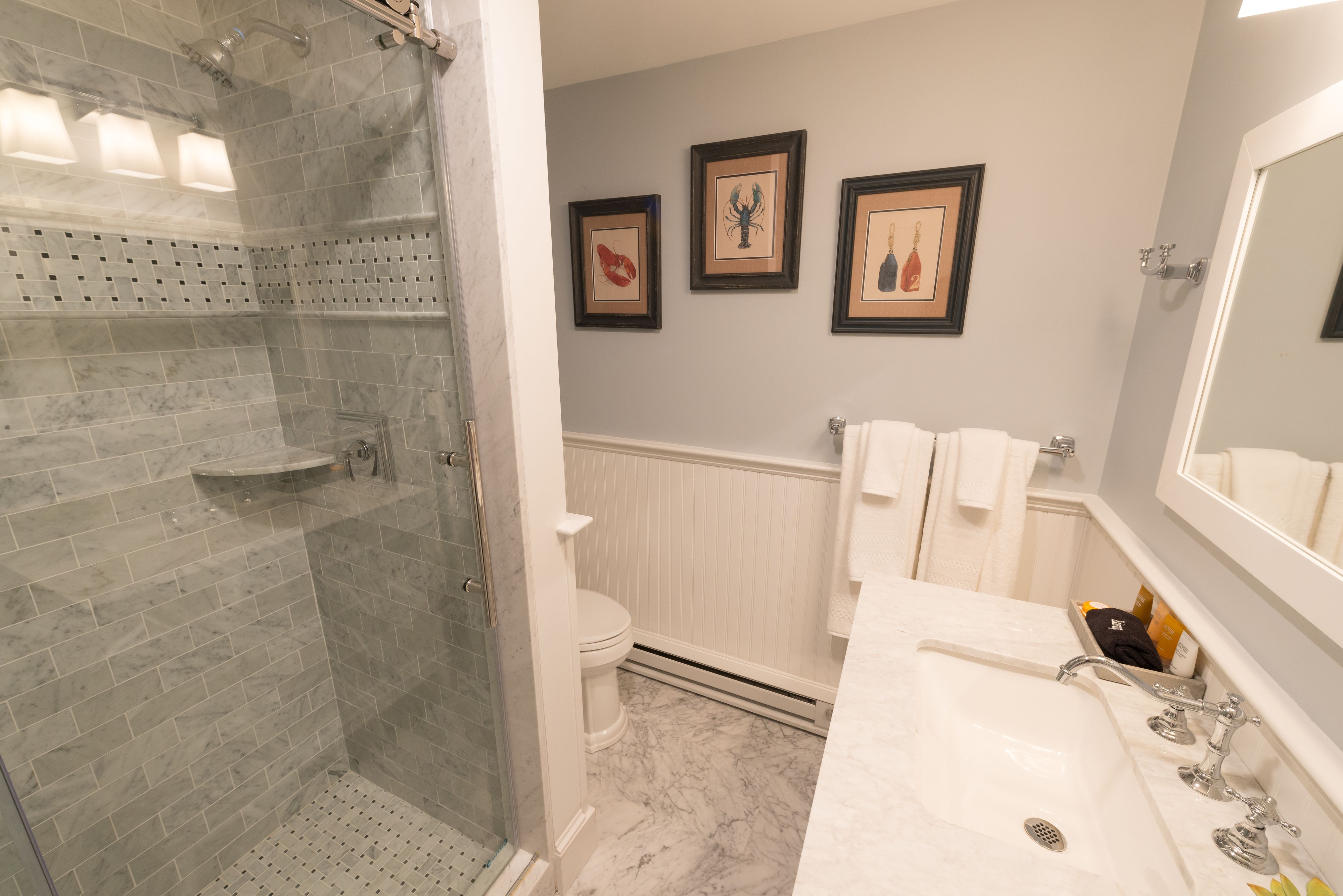 A bathroom with a glass-enclosed shower, a white toilet, a sink with a mirror, and three framed pictures on the wall.