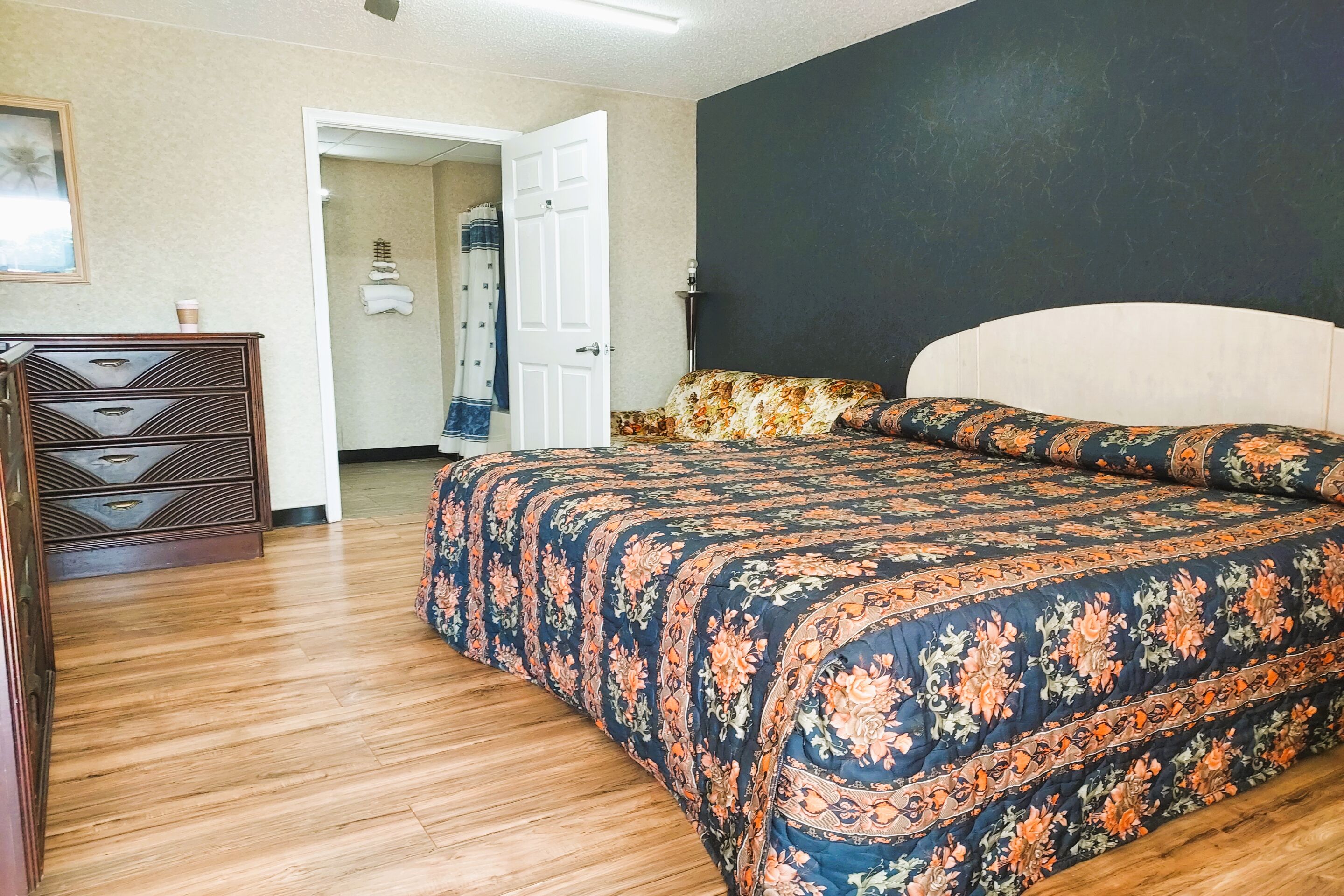Una camera da letto con pavimento in legno, un letto con copripiumino floreale, una cassettiera e una porta che conduce in un'altra stanza.