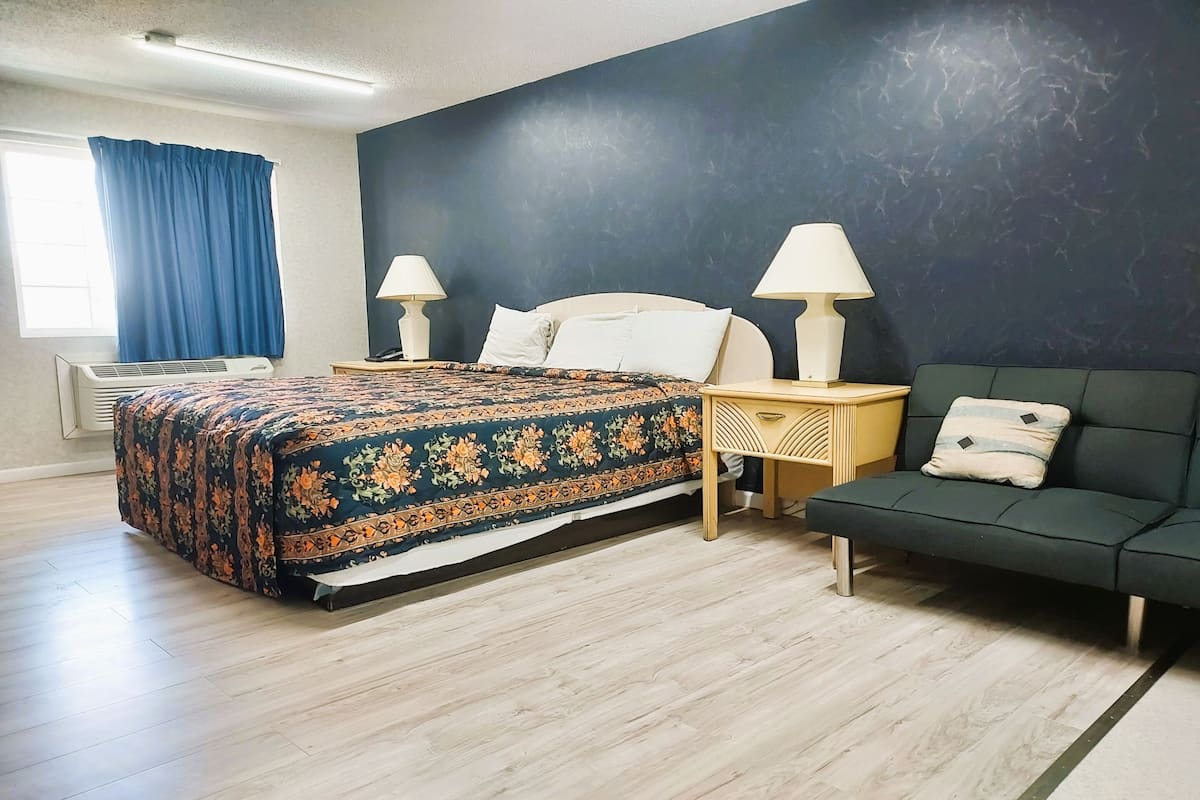 A hotel room with a bed, bedside lamps, a sofa, a window with curtains, and an air conditioning unit.