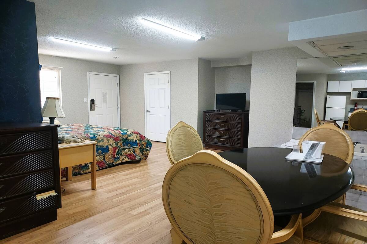 A room with a round dining table, chairs, a bed with a colorful blanket, a dresser, a television, and a kitchen area with cabinets and appliances.