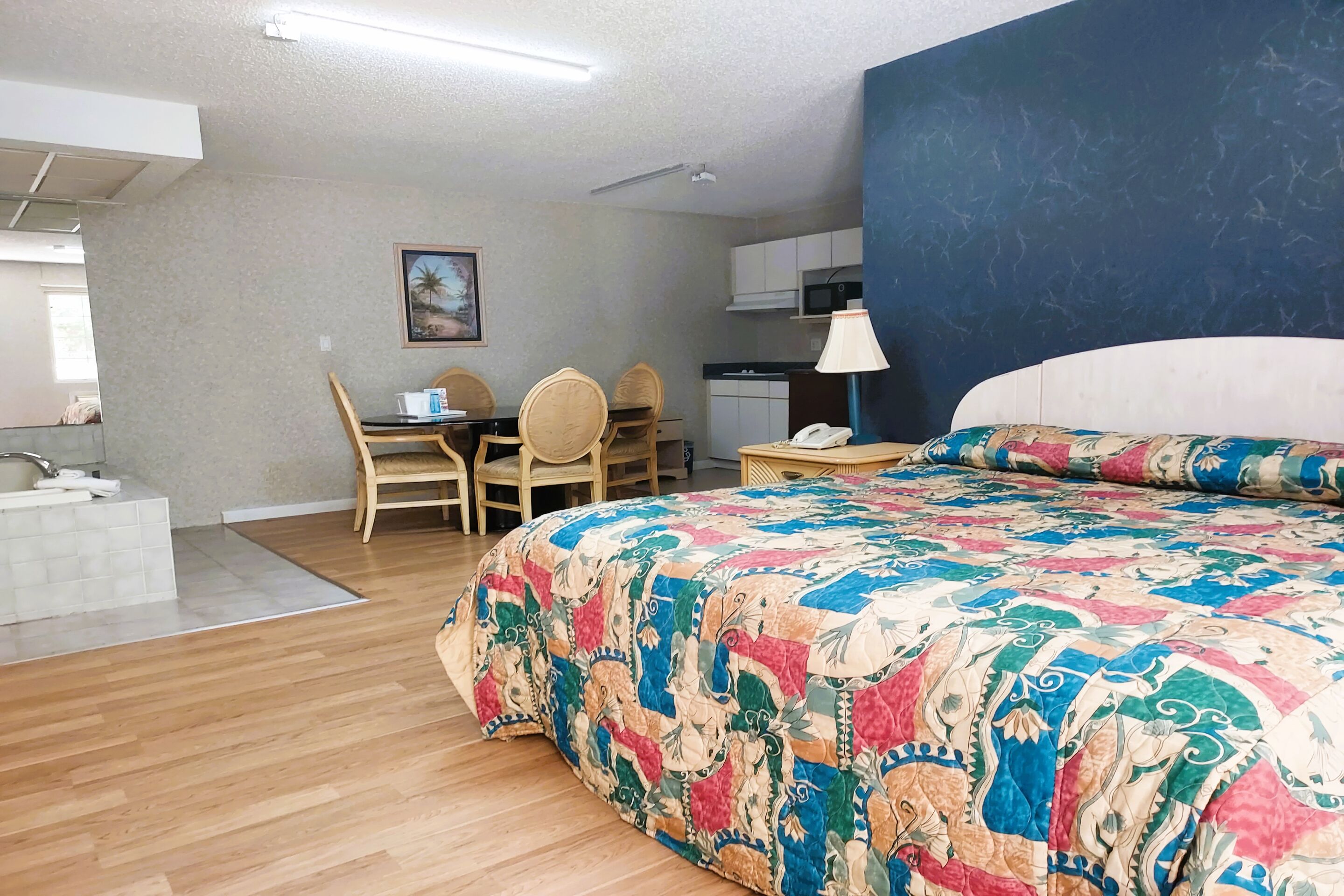 Quarto de hotel com uma cama grande, uma escrivaninha com cadeiras, uma pequena cozinha e um banheiro com pia e espelho.