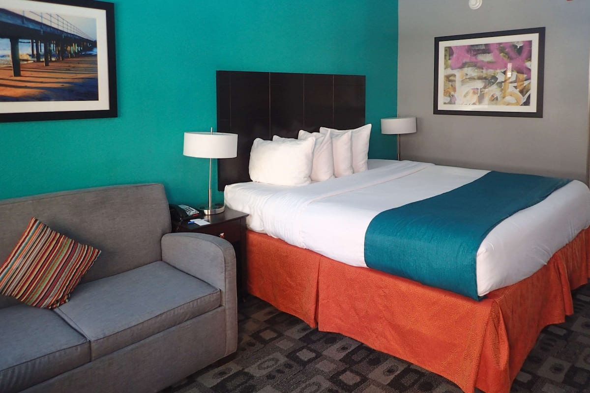 A hotel room with a bed, a sofa, a nightstand, a lamp, and two framed pictures on the walls.