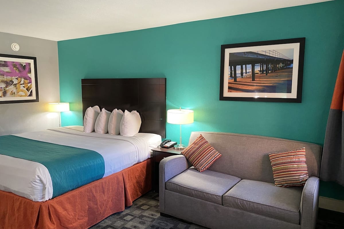 A hotel room with a bed, a sofa, two lamps, a framed picture, and a patterned rug.