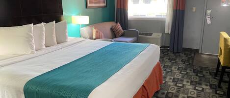 A hotel room with a large bed, a desk, a chair, a window with curtains, and a wall-mounted air conditioner.
