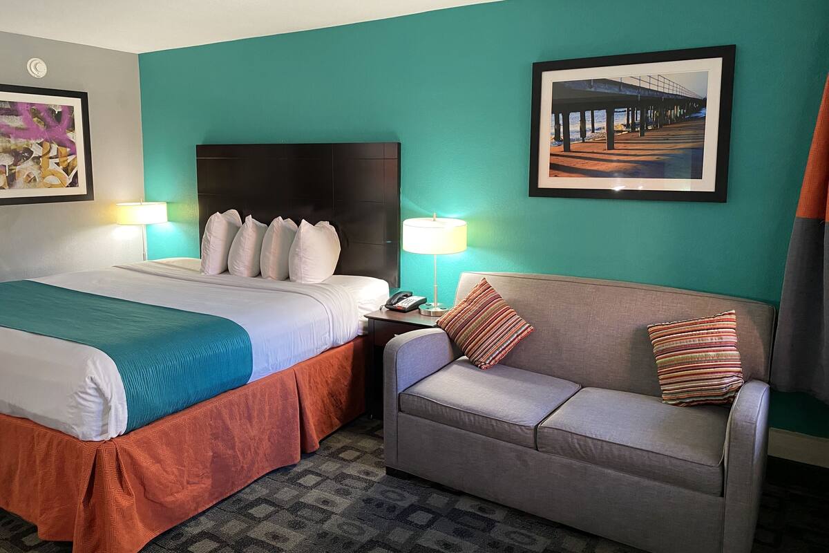 A hotel room with a bed, a sofa, two lamps, a phone on the nightstand, and a framed picture on the wall.