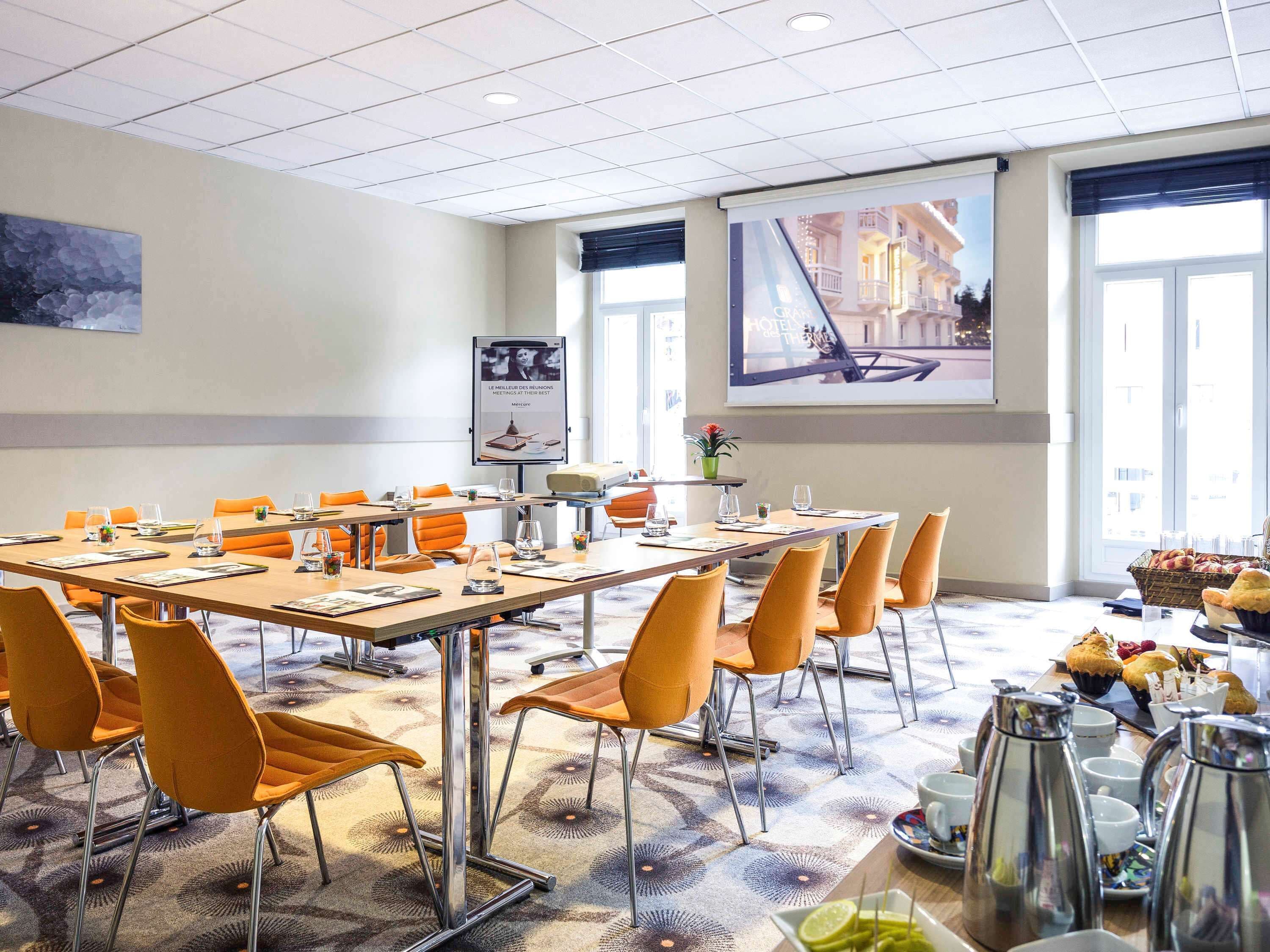 Ein Konferenzraum mit einem langen Tisch, orangefarbenen Stühlen, einer Projektionsleinwand und einem großen Fenster.