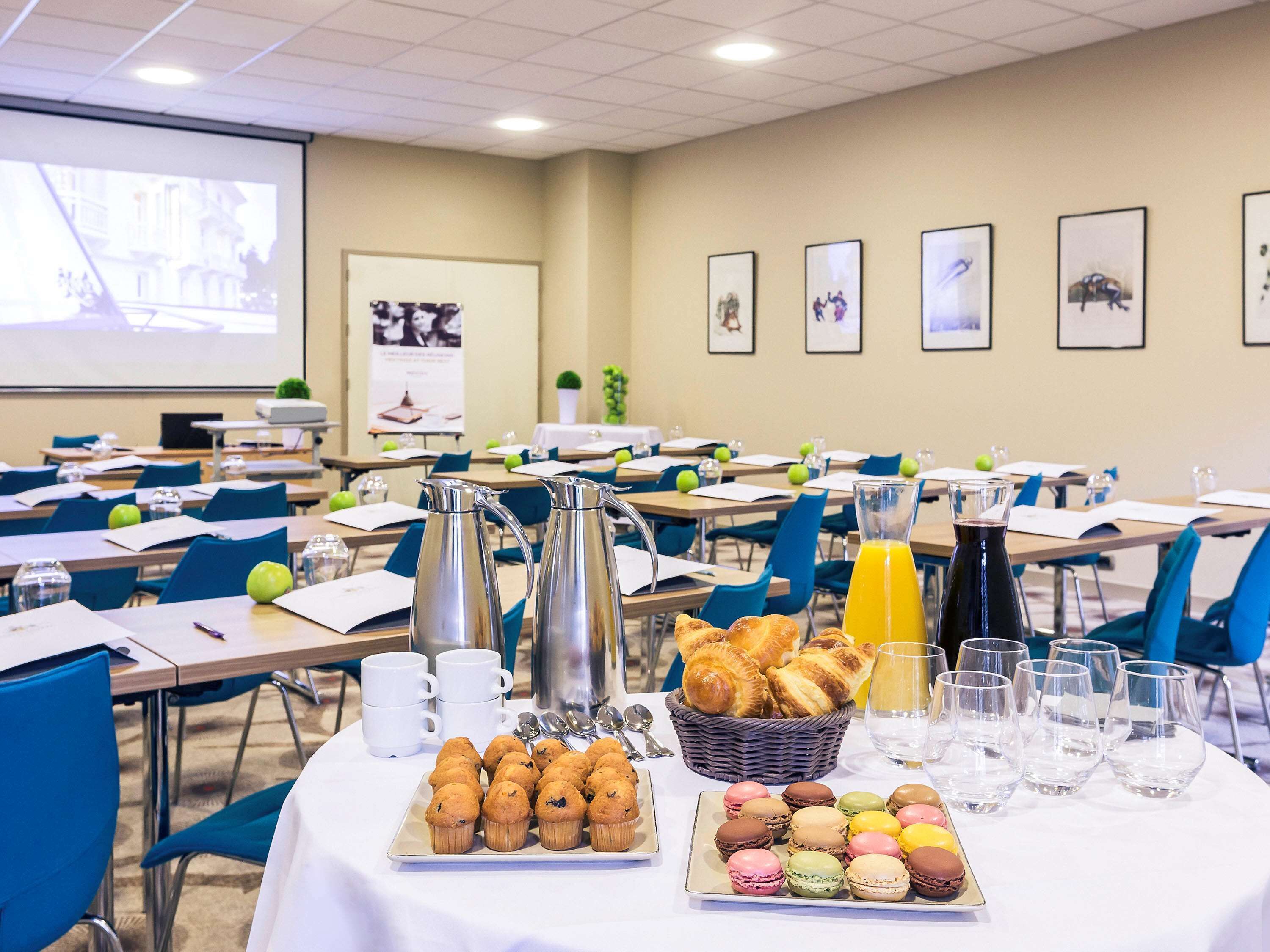 Une salle de conférence avec des tables disposées pour une réunion, équipée de cafetières, de verres à eau et d’un distributeur de jus.