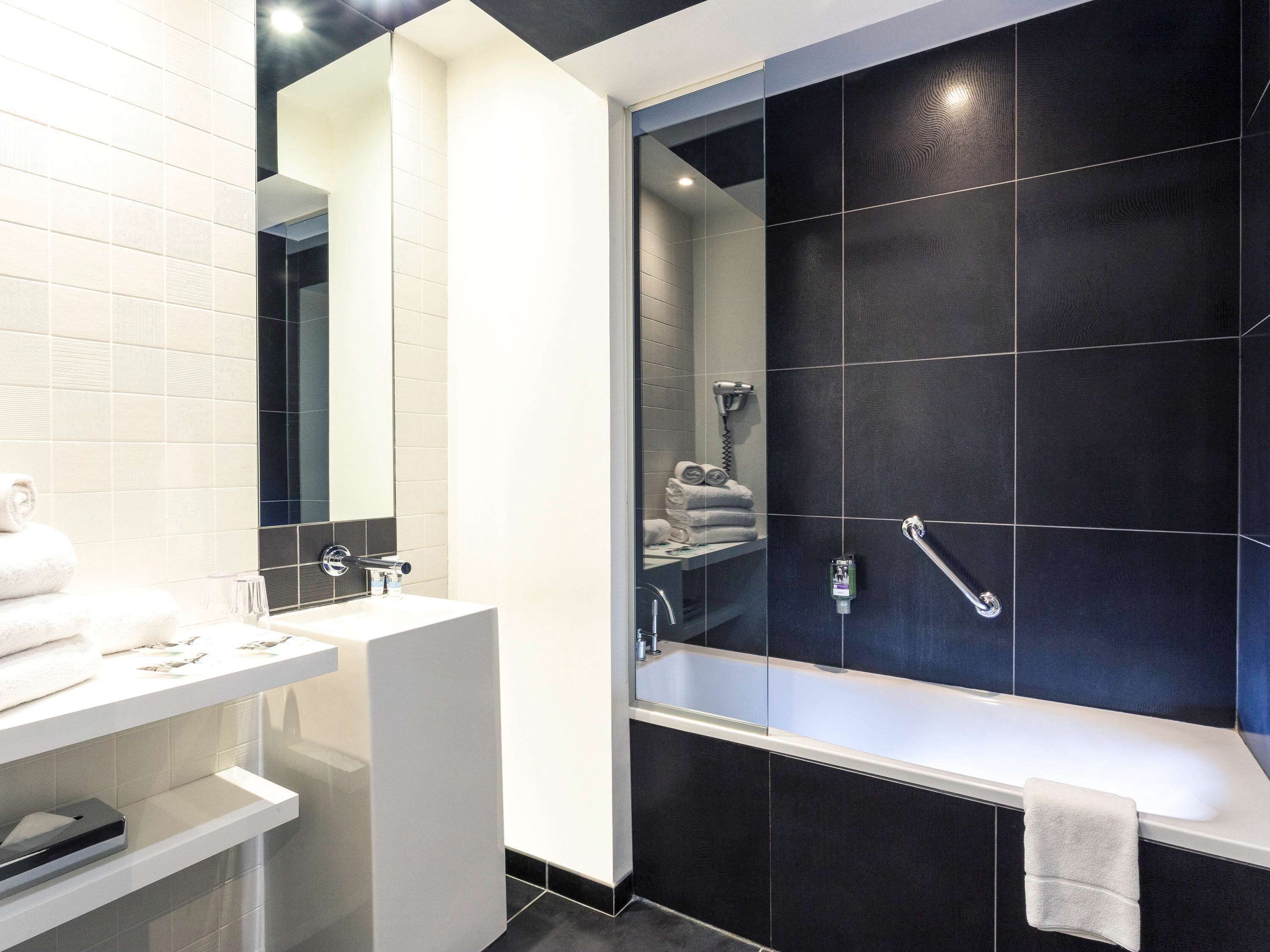 Un baño moderno con tocador blanco, un espejo grande, zona de ducha con azulejos oscuros y una toalla blanca en el suelo.