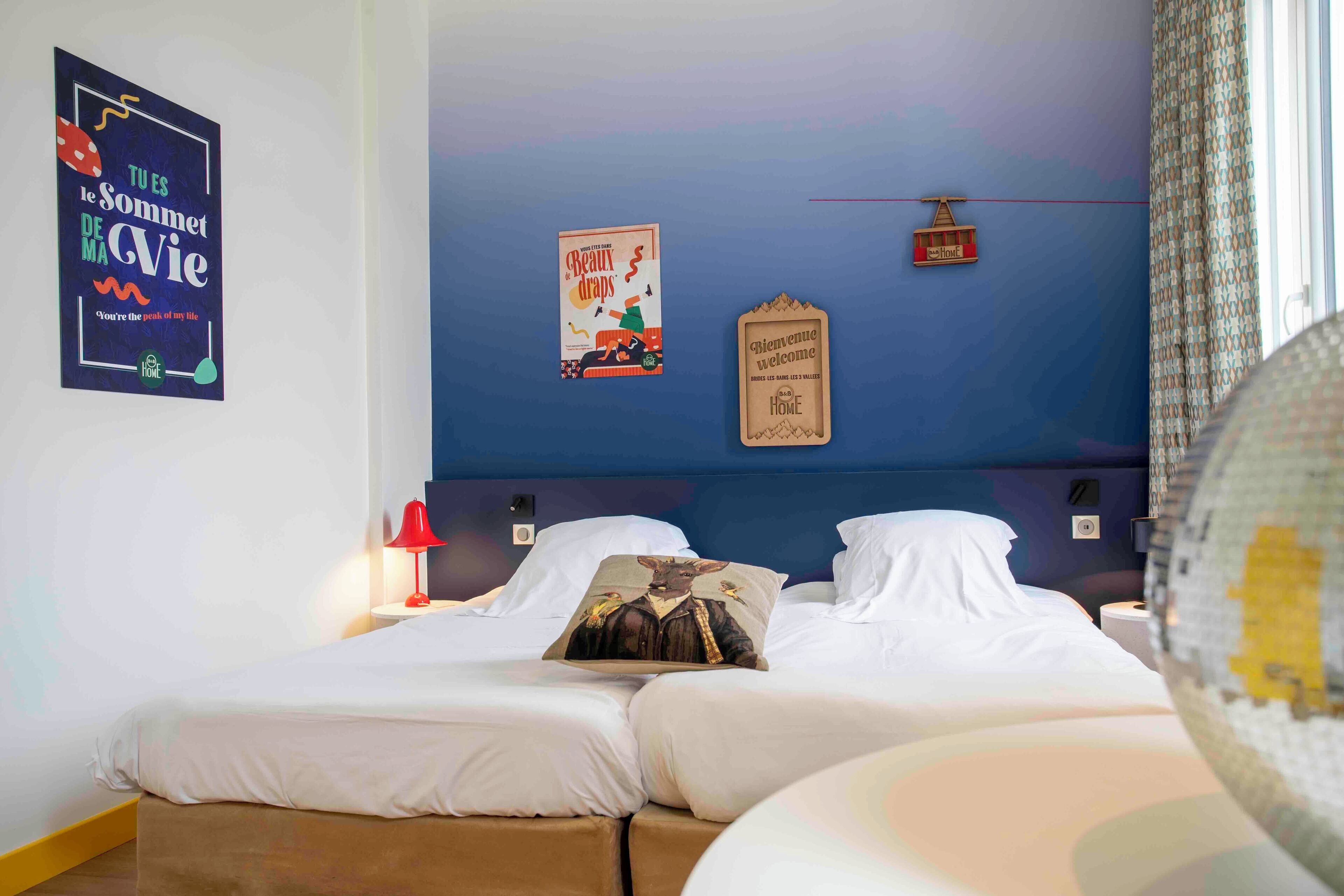 Quarto de hotel com cama, mesa de cabeceira, abajur vermelho e decorações na parede, incluindo um cartaz e uma placa de boas-vindas.