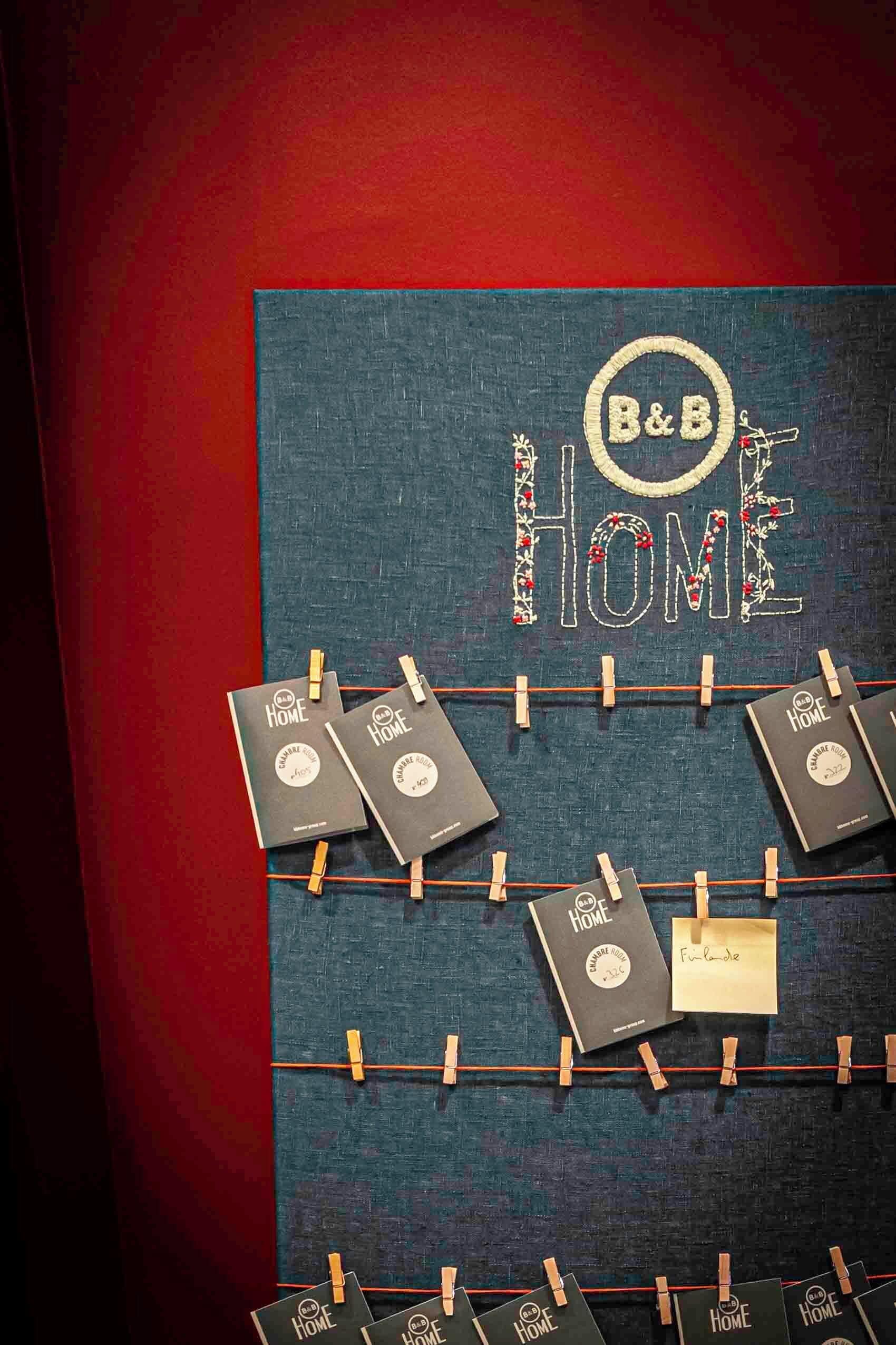 A wall with 'B&B HOME' embroidered in a decorative style, featuring several black notebooks clipped to the wall with wooden clothespins.