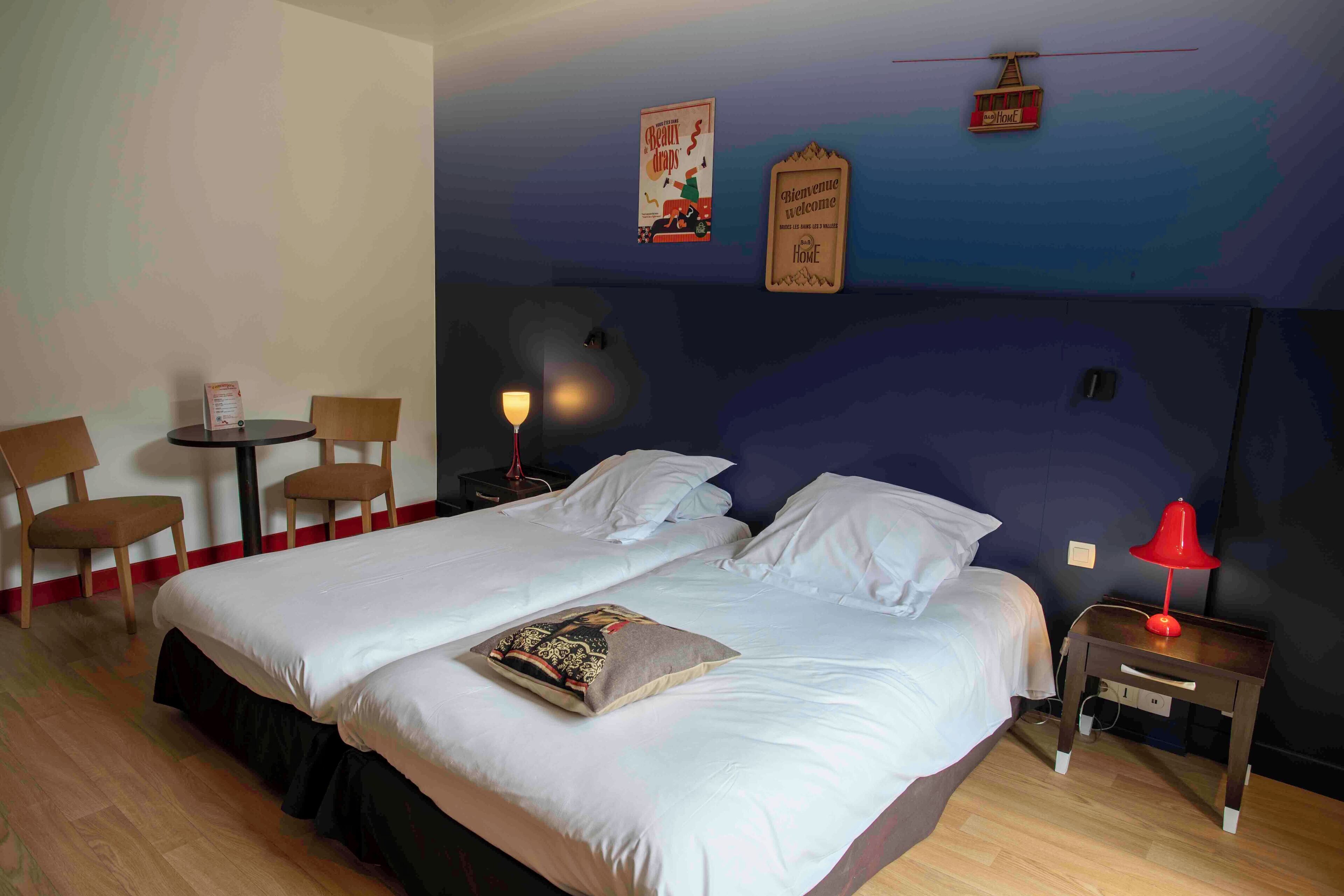 Habitación de hotel con una cama grande, dos sillas, una mesita pequeña y una mesita de noche con lámpara.