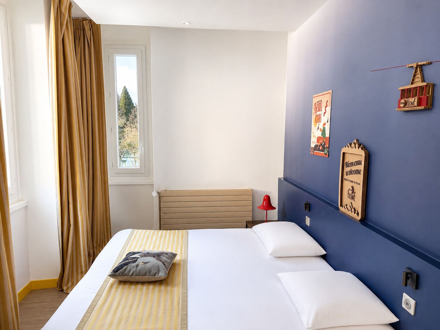 Une chambre d’hôtel avec un lit, une fenêtre avec des rideaux, une lampe fixée au mur et un panneau d’accueil.
