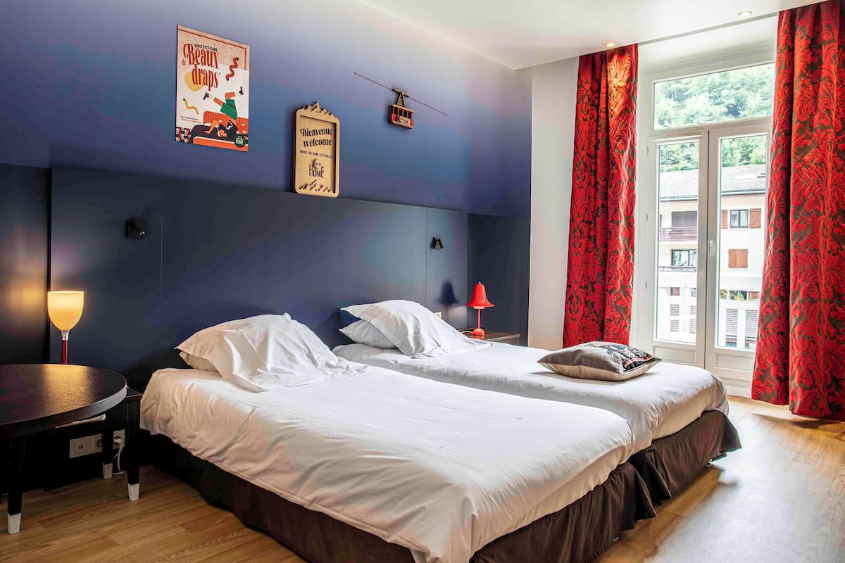 Habitación de hotel con dos camas, una mesita, una lámpara, un cartel en la pared y una ventana con cortinas.