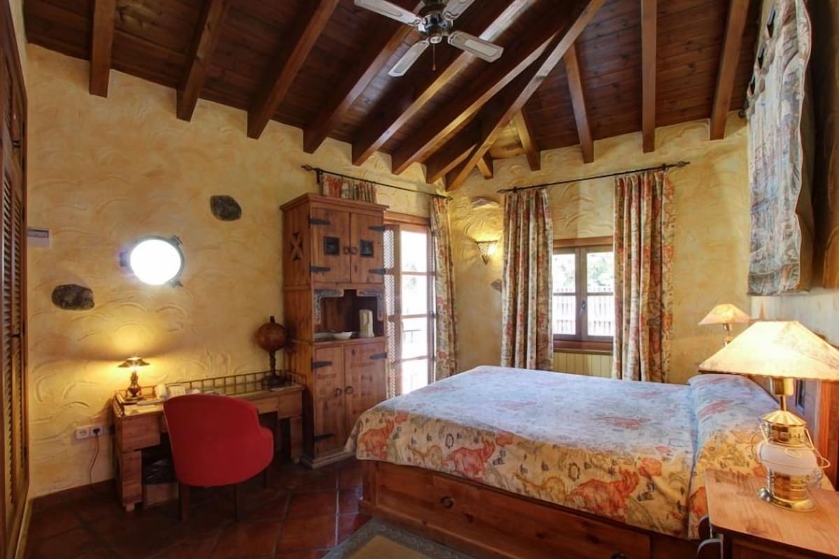 Un dormitorio con techo de madera, una cama con colcha estampada, un escritorio con lámpara y una silla roja.
