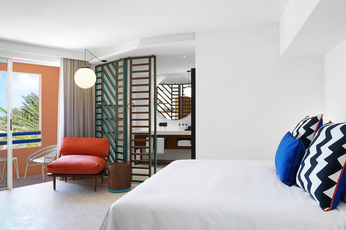 A modern hotel room with a bed, a red chair, a wooden side table, a large window with a view of palm trees, and a balcony.