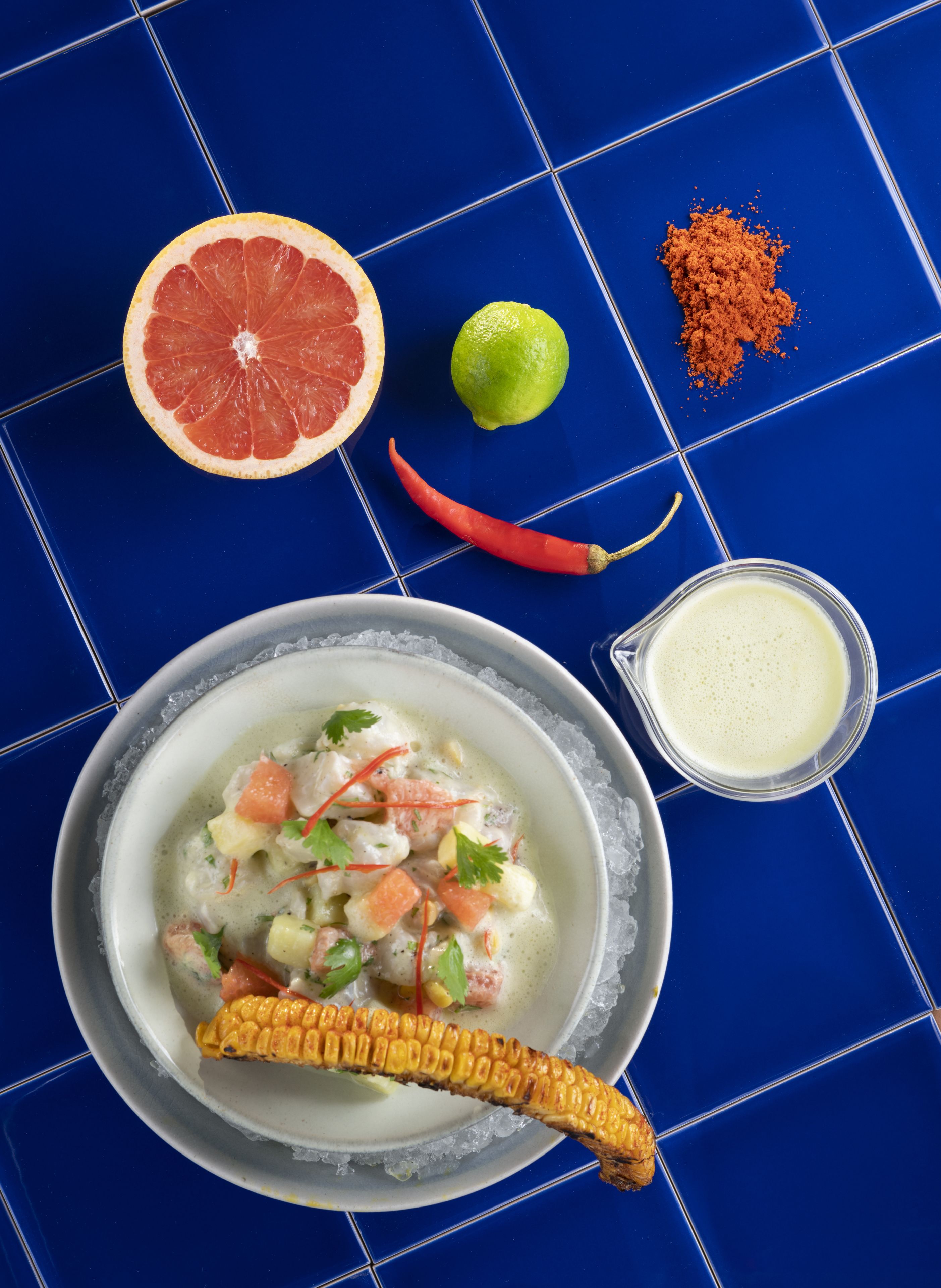 A bowl of soup with corn on the cob, a half grapefruit, a lime, and a red chili pepper on a blue tiled surface.