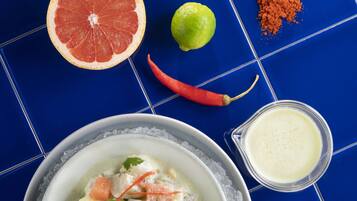 A bowl of soup with corn on the cob, a half grapefruit, a lime, and a red chili pepper on a blue tiled surface.