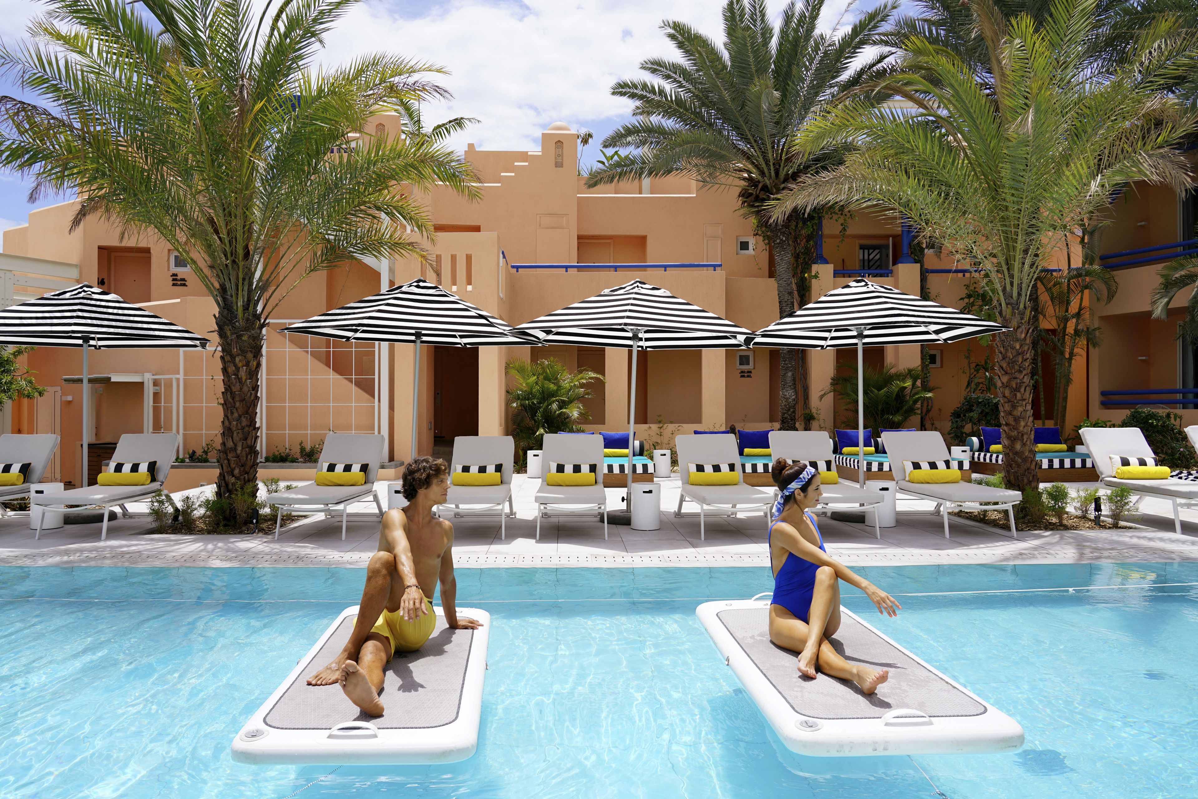 Zona de piscina con sillas de descanso y sombrillas, rodeada de palmeras y un edificio con balcones.