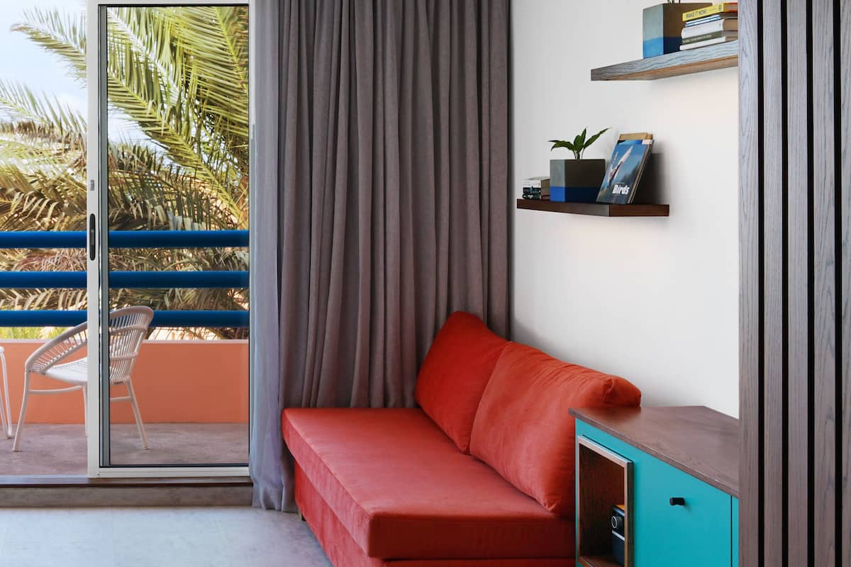 A modern living room with a red sofa, a teal cabinet, and a sliding glass door leading to a balcony with a view of palm trees.