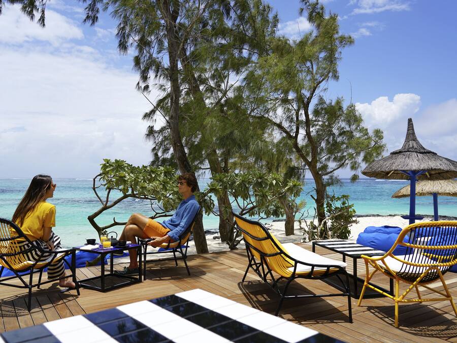 Una terraza de madera con asientos exteriores, con vista a una playa de aguas azules y un paraguas de paja.