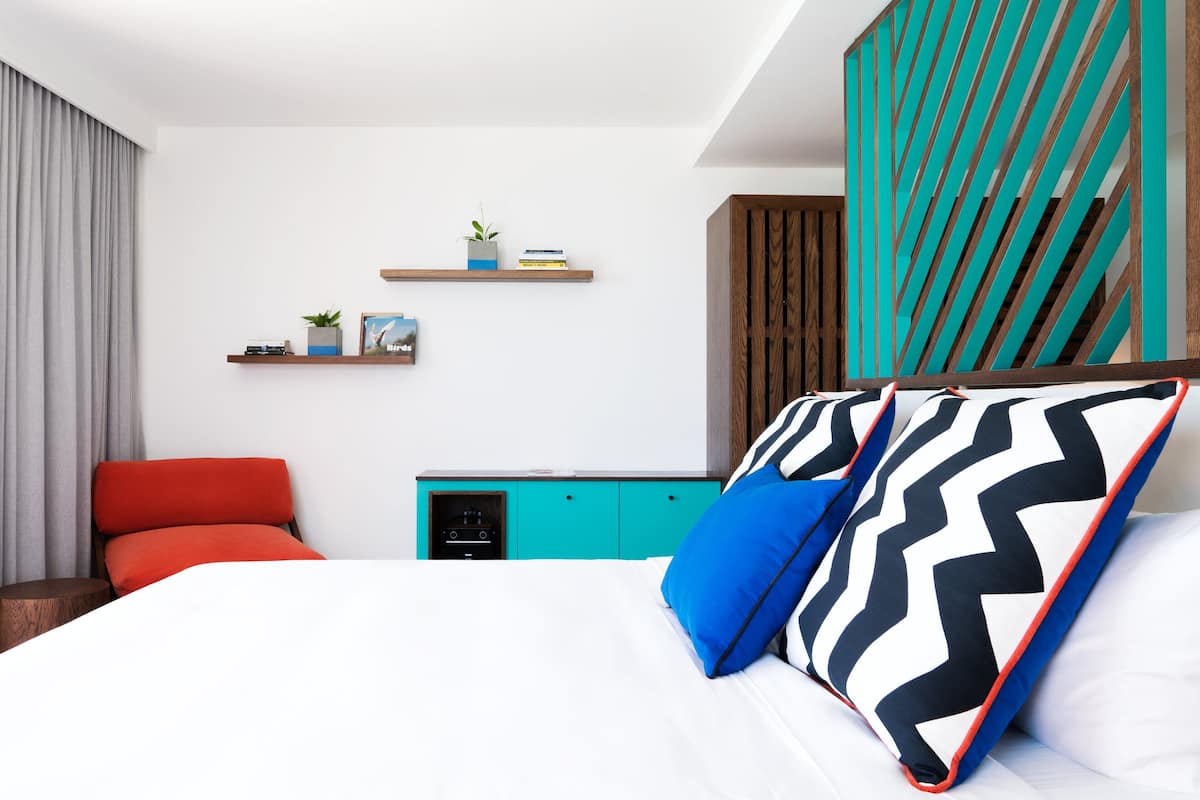 A modern hotel room with a bed, a red chair, a teal dresser, and a wall-mounted shelf with books and a plant.