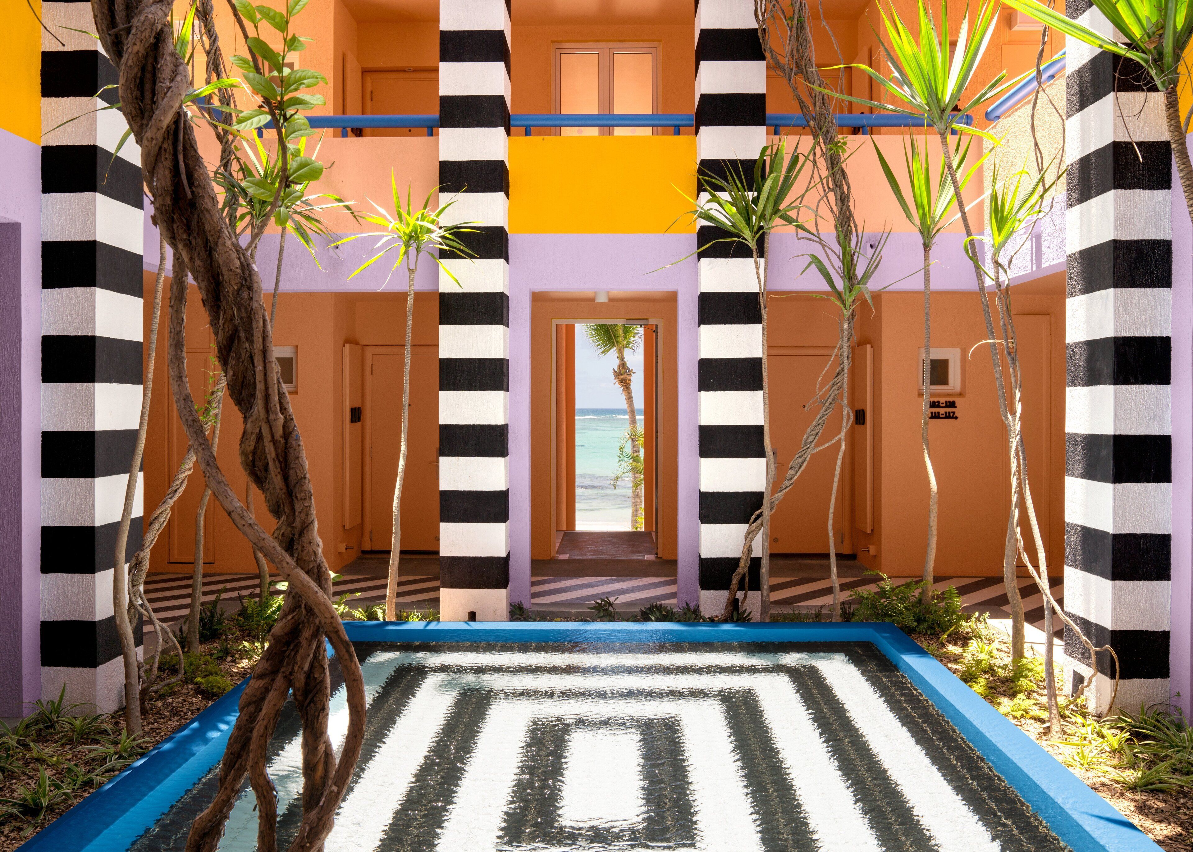 A hotel lobby with a checkered floor pattern, palm trees, and a view of the ocean through an open door.