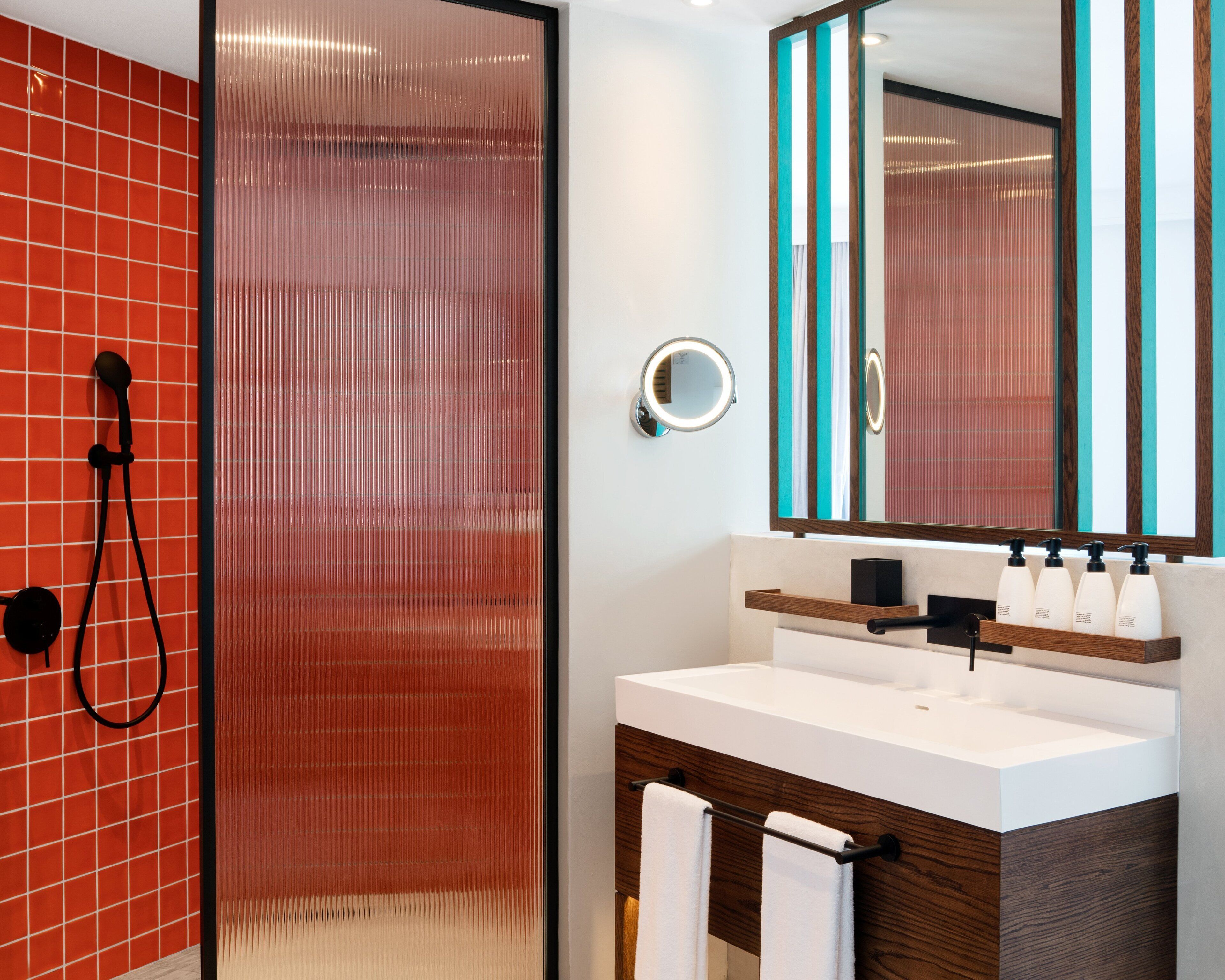 Un baño moderno con cabina de ducha de vidrio, un lavabo blanco con mueble de madera y un espejo con detalles azul turquesa.