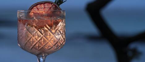A cocktail glass with a decorative pattern, garnished with a sprig and a slice, placed on a table with a blurred background of a tree and sky.