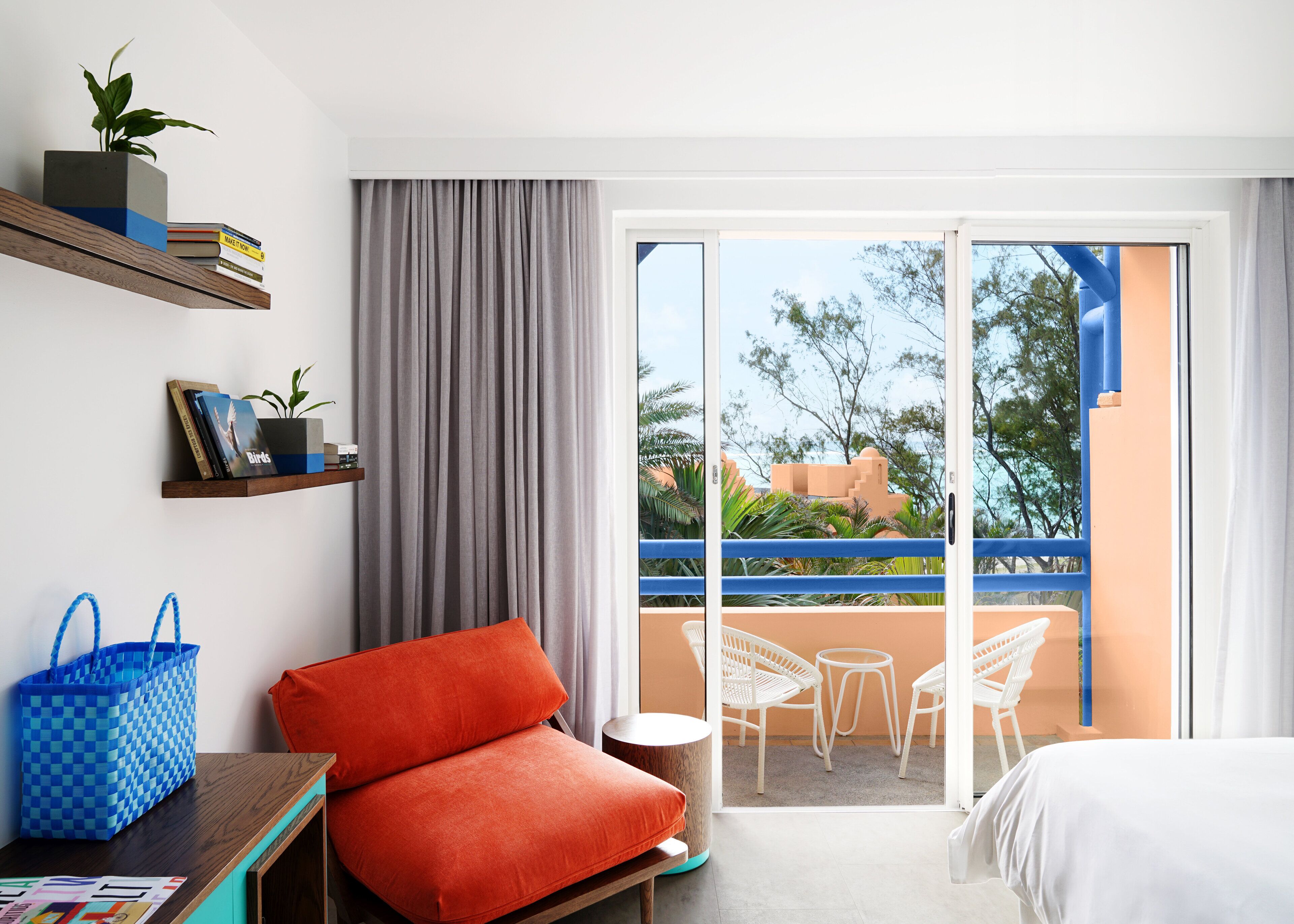A hotel room with a balcony, a red sofa, a small table, a blue checkered bag, and a plant on a shelf.