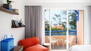 A hotel room with a balcony, a red sofa, a small table, a blue checkered bag, and a plant on a shelf.