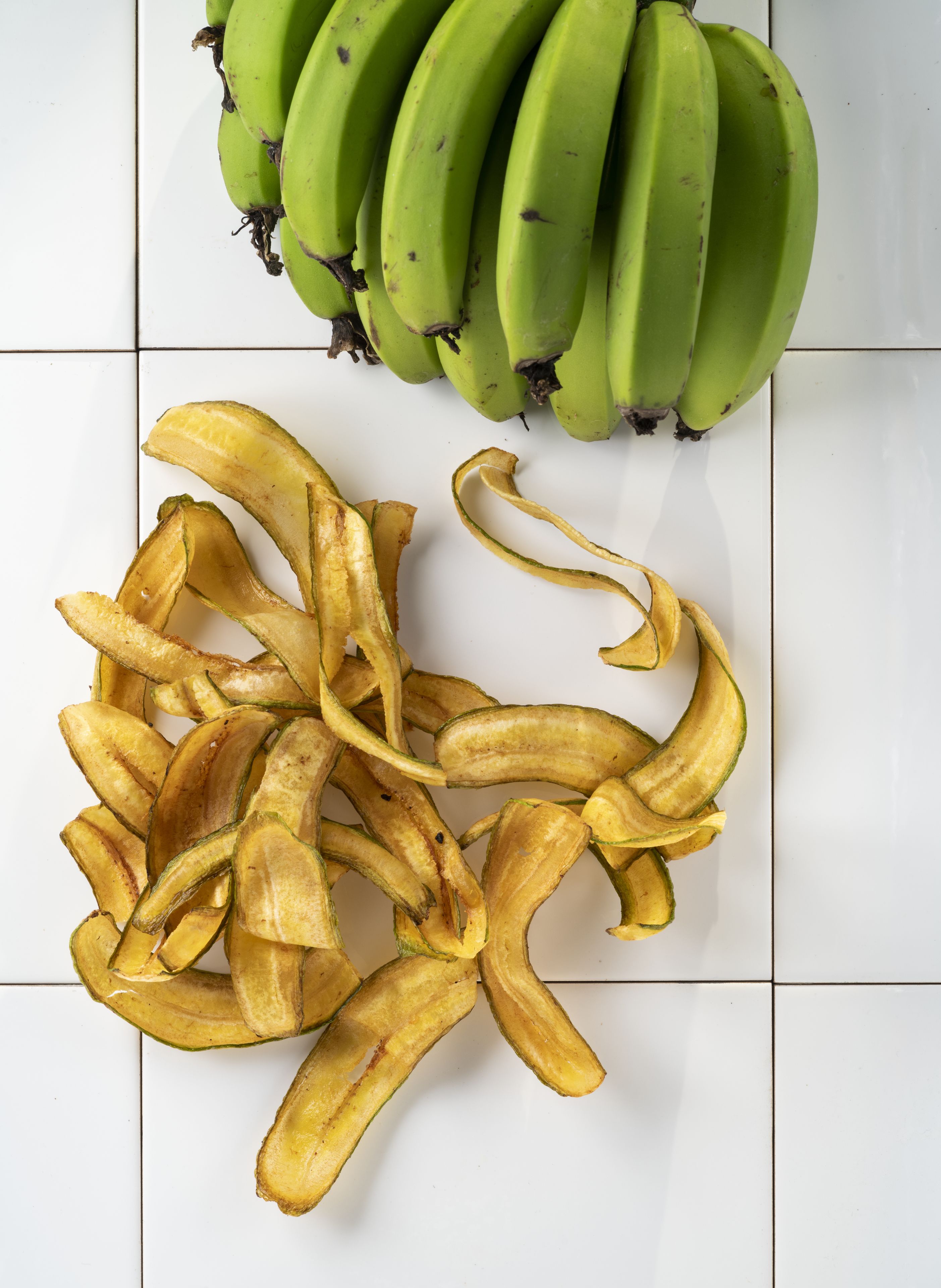 A bunch of green bananas and a pile of banana peels on a tiled surface.