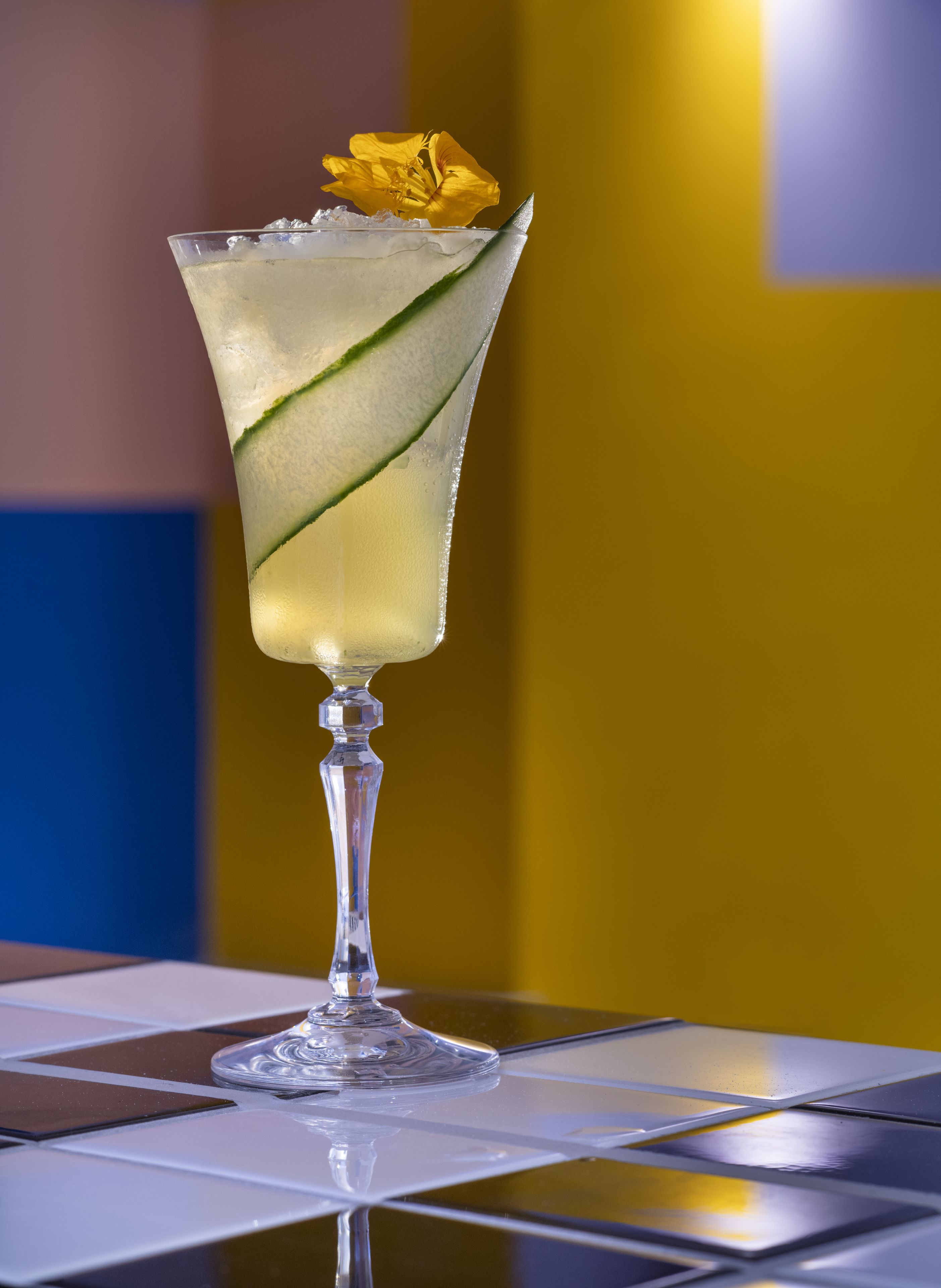 Un cocktail guarnito con cetriolo e un fiore giallo su un bicchiere, posizionato su un tavolo con superficie piastrellata.