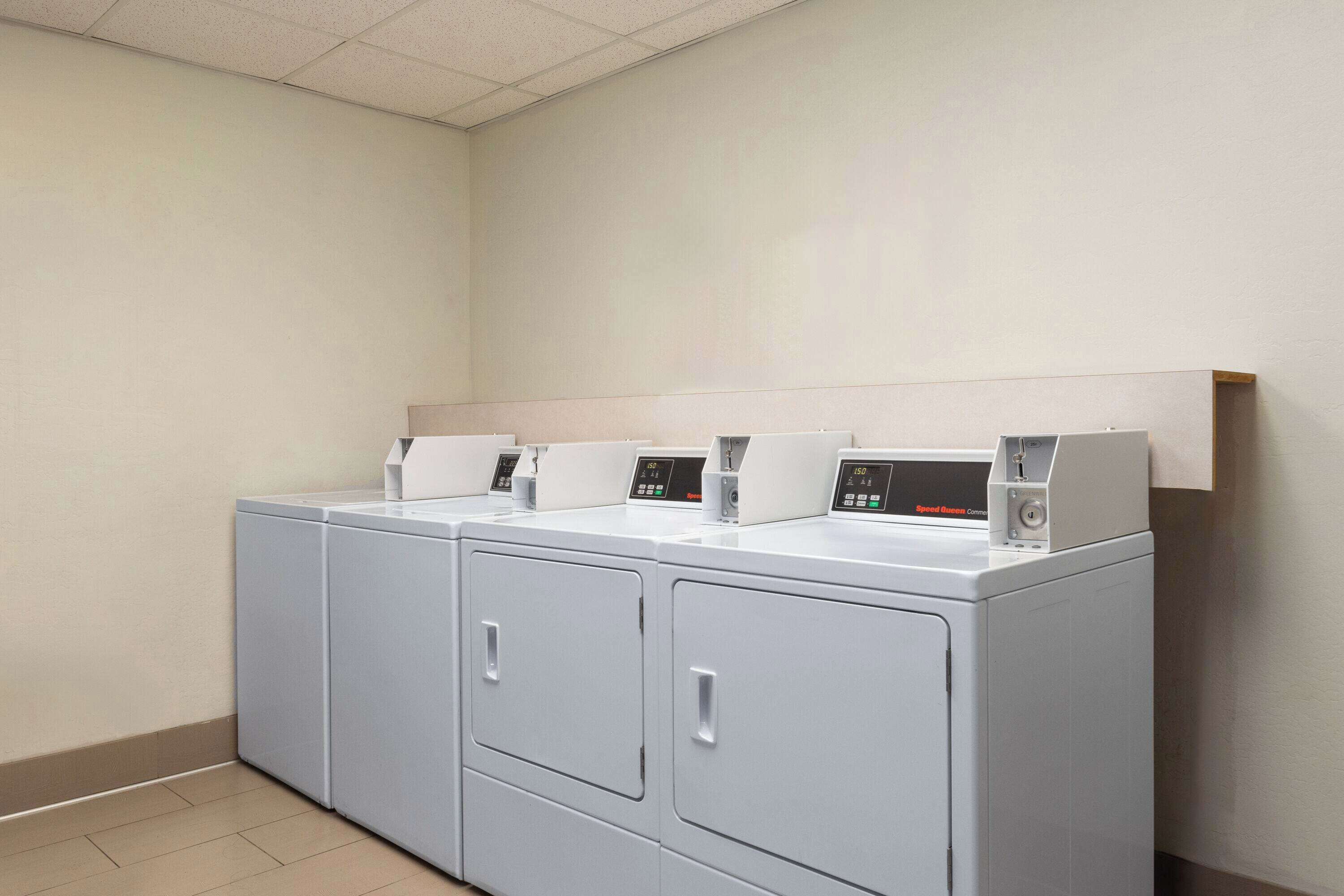 A row of white, modern, front-loading washers and dryers in a clean, tiled room with a plain wall and a wooden shelf above the dryers.
