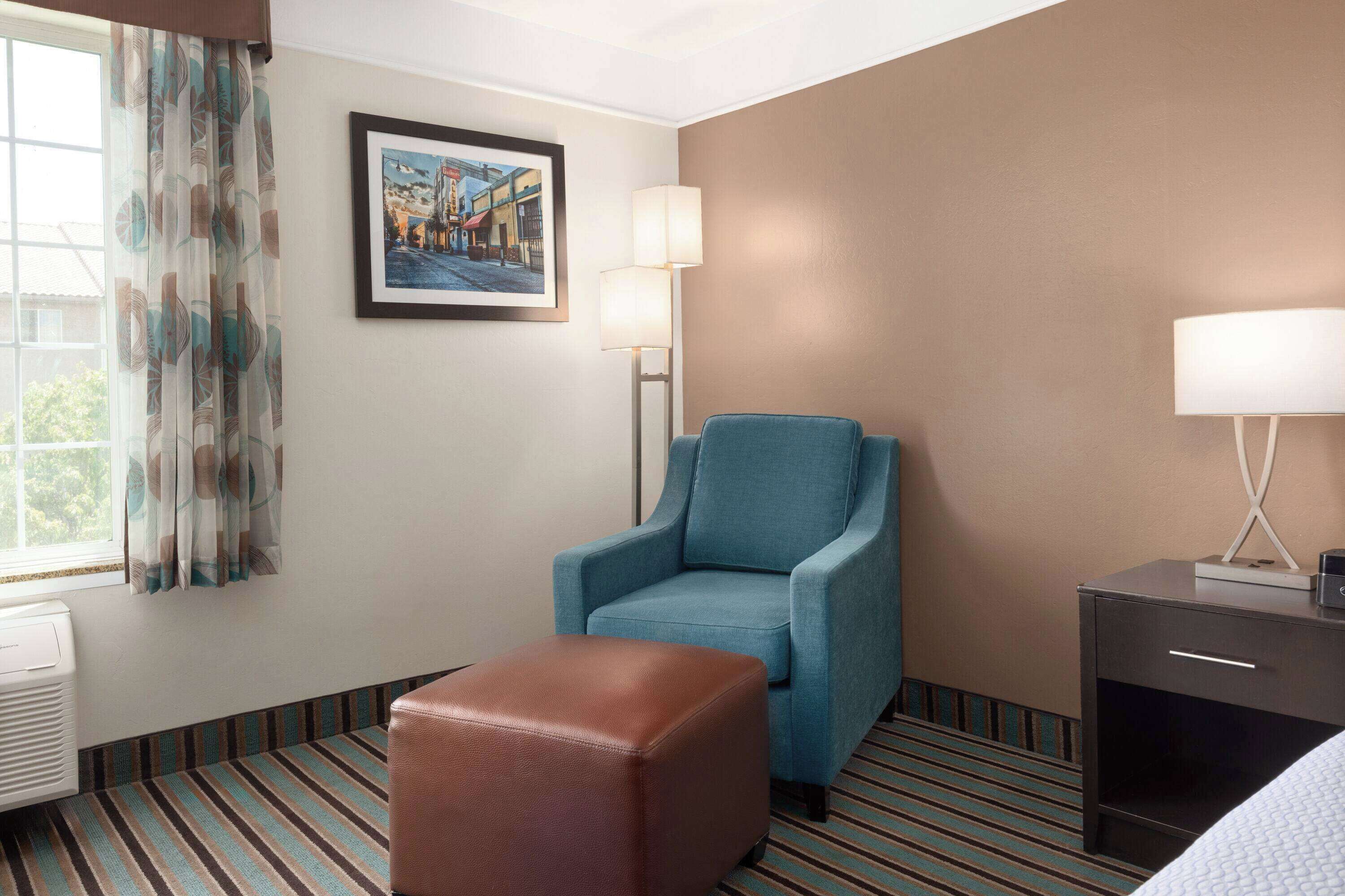 A hotel room with a striped carpet, a blue armchair, a brown ottoman, a bedside table with a lamp, and a framed picture on the wall.