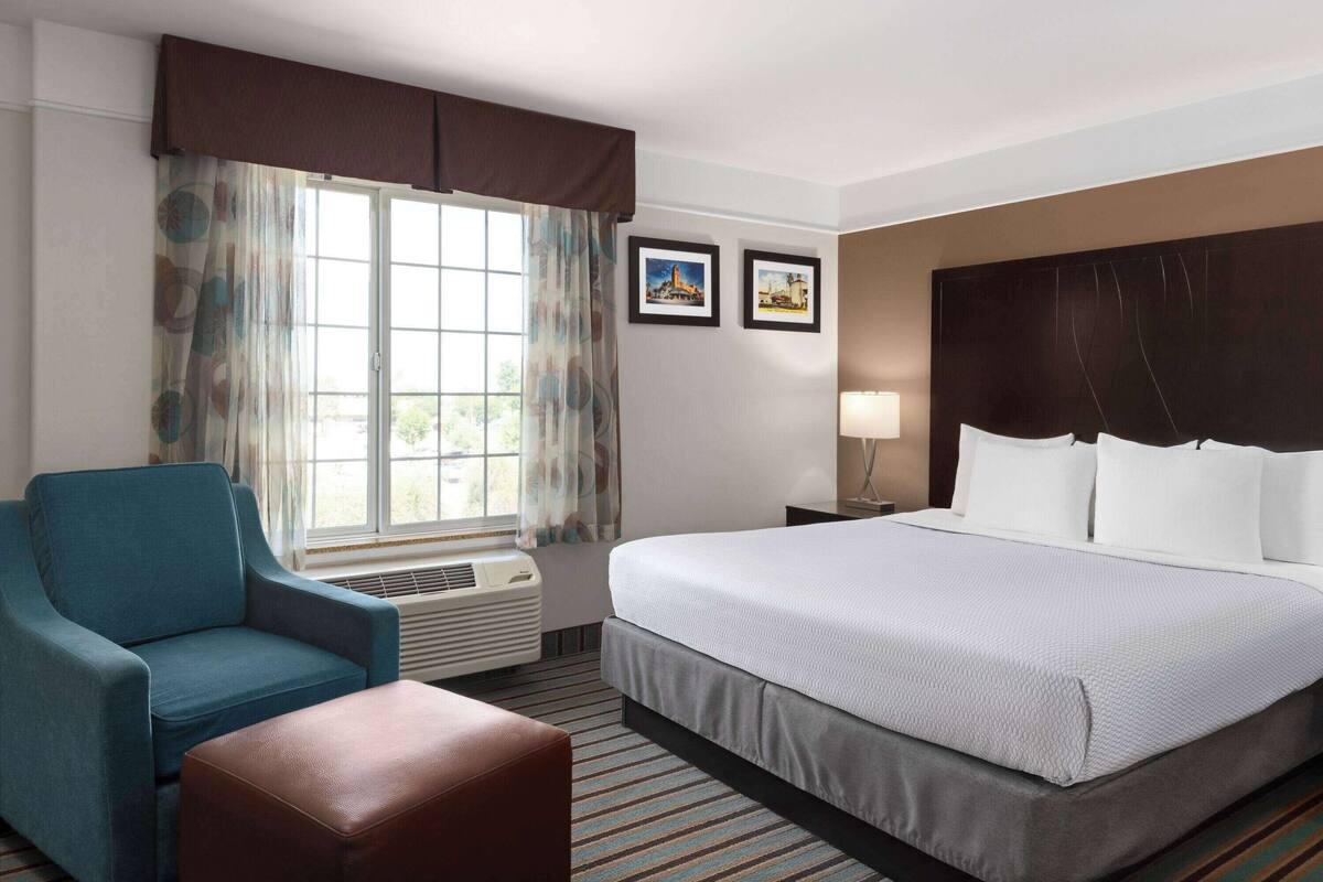 A hotel room with a large bed, a blue armchair, a brown ottoman, a window with curtains, and two framed pictures on the wall.