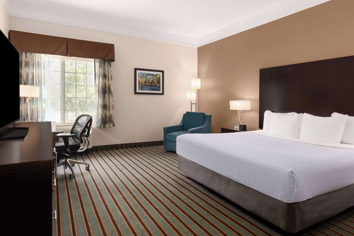 A hotel room with a large bed, a desk with a chair, a window with curtains, and a painting on the wall.