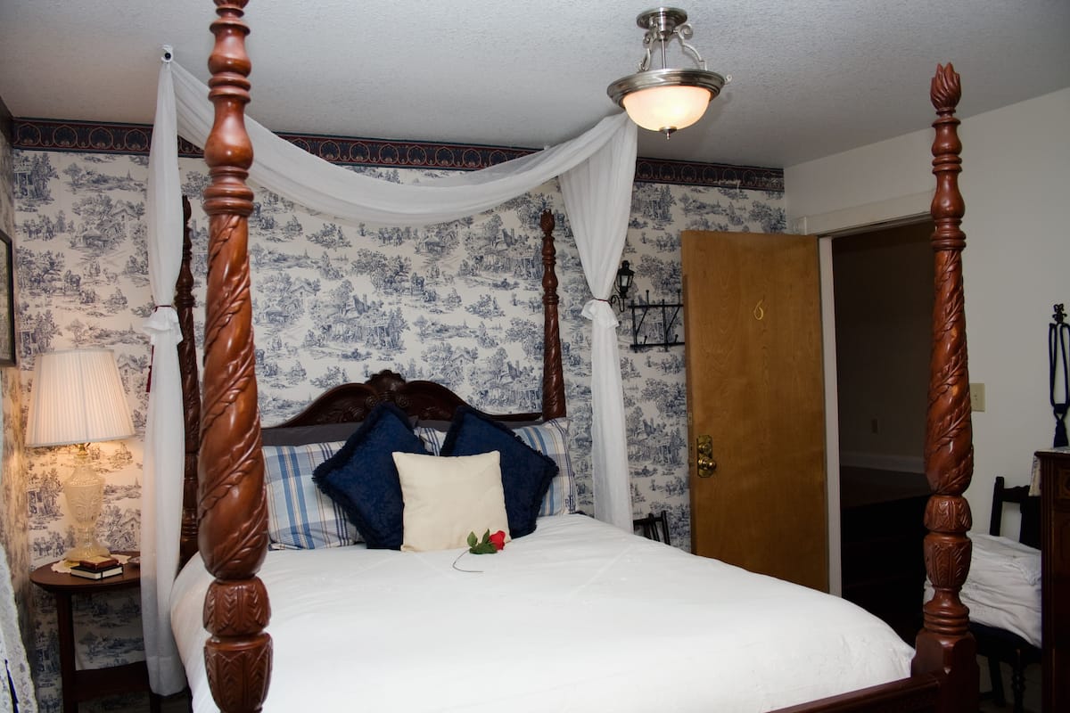 A four-poster bed with a canopy, a nightstand with a lamp, and a wall with blue floral wallpaper.