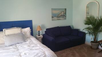 A bedroom with a bed, a blue headboard, a blue sofa, a lamp, a painting, and a potted plant.