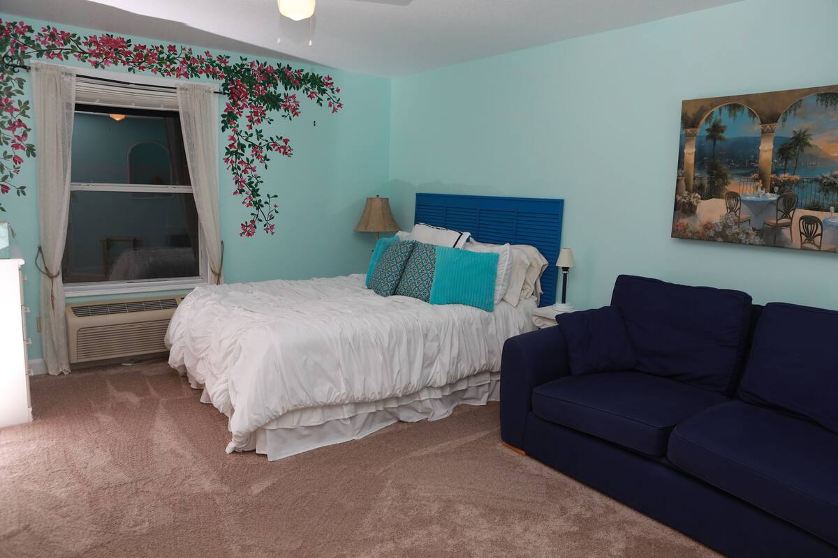 A bedroom with a bed, a blue headboard, a dark blue sofa, a window with curtains, and a wall painting.