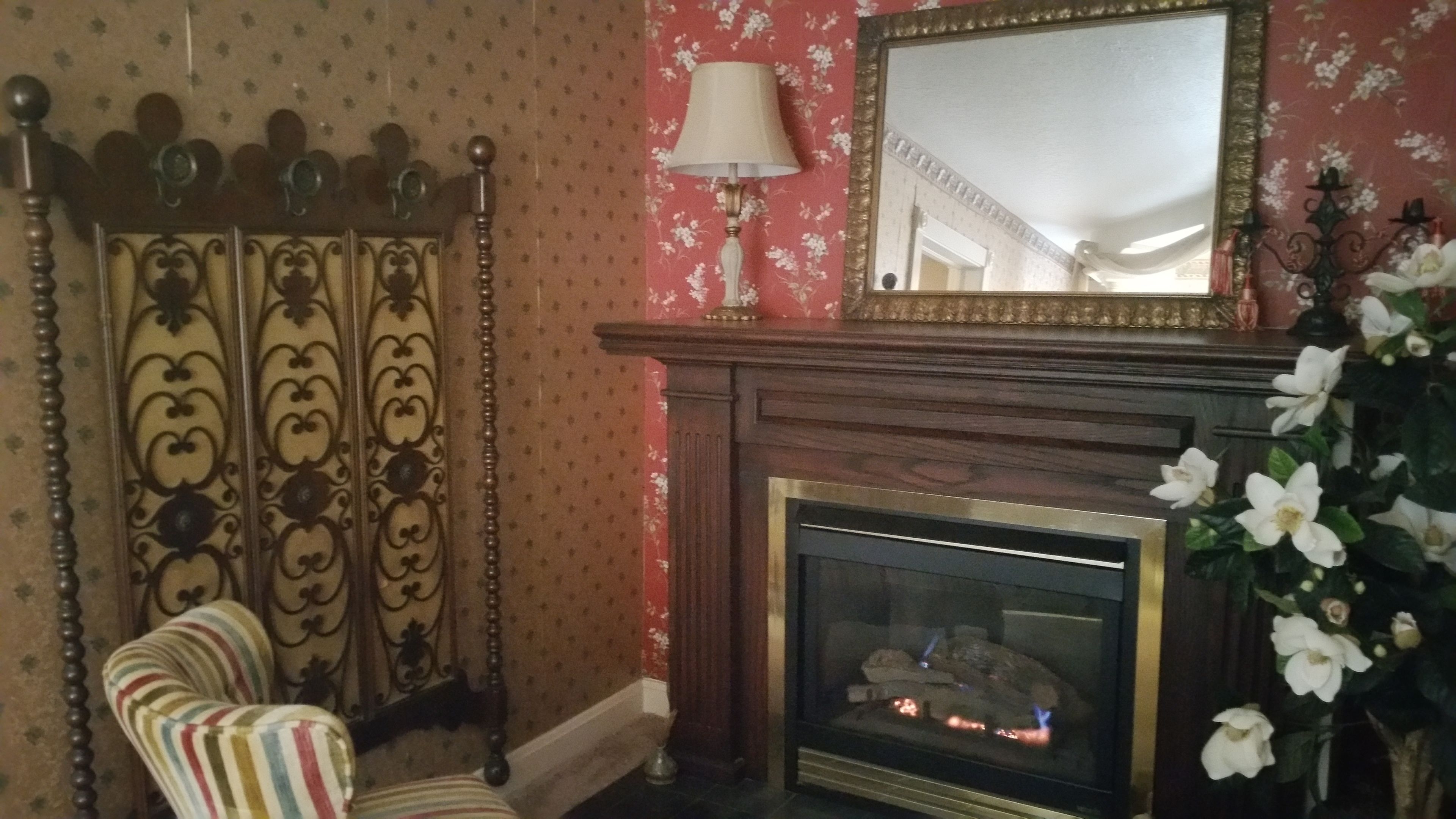 A room with a patterned wallpaper, a fireplace with a mirror above it, a striped armchair, and a vase with white flowers.