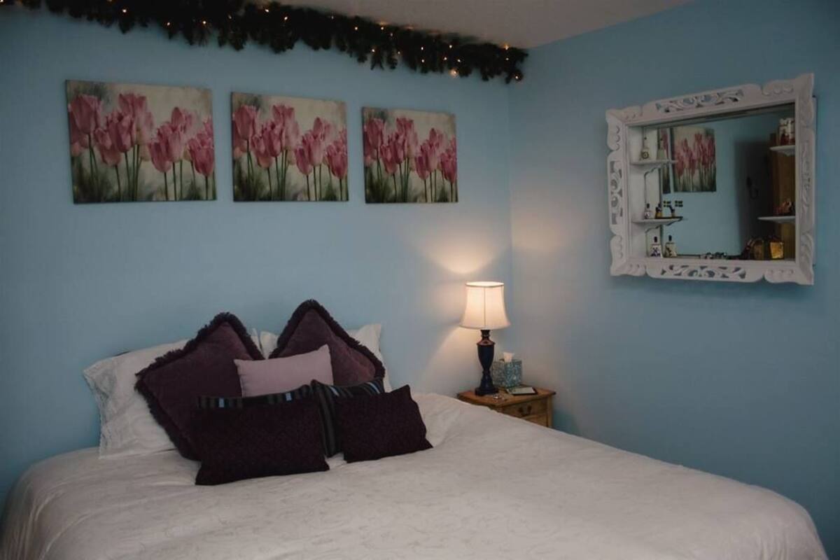 A bedroom with a bed, a nightstand with a lamp, and three wall-mounted paintings of pink flowers.