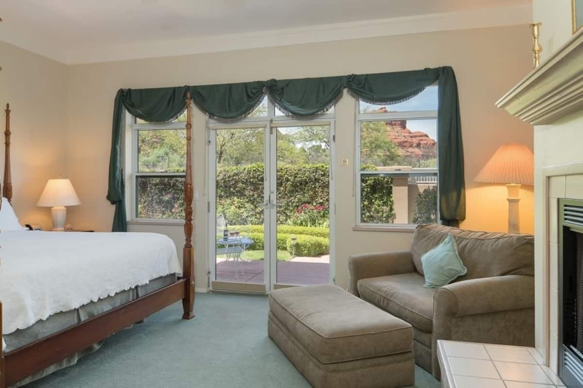 A bedroom with a four-poster bed, a sofa, and an ottoman. There is a fireplace and a view of a garden through a glass door.